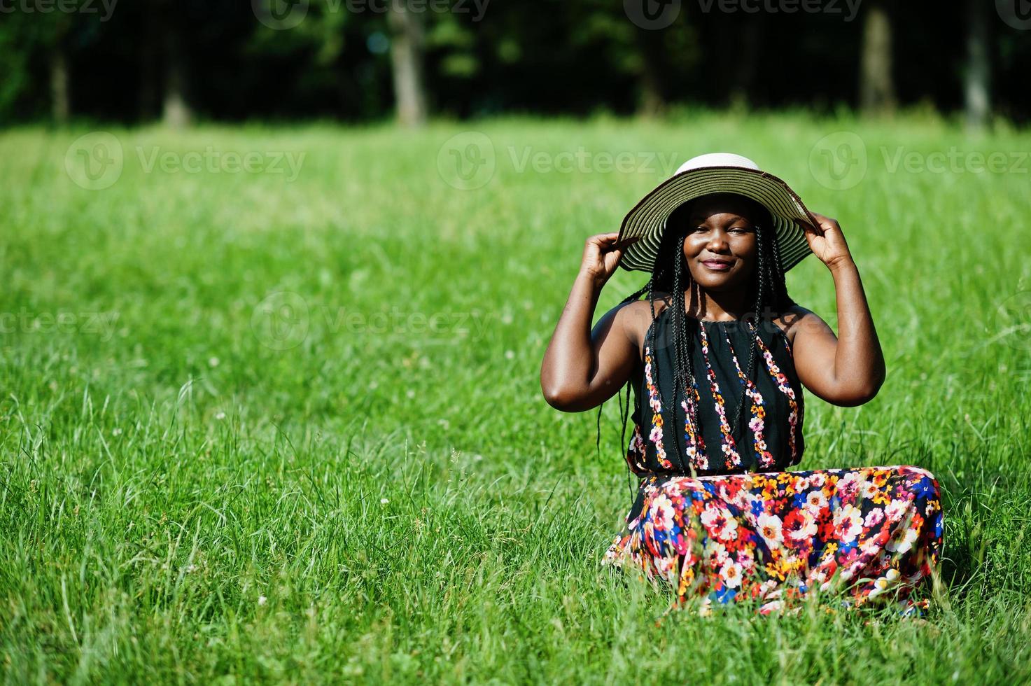 retrato de una hermosa mujer afroamericana de 20 años con sombrero de verano sentada en la hierba verde en el parque. foto