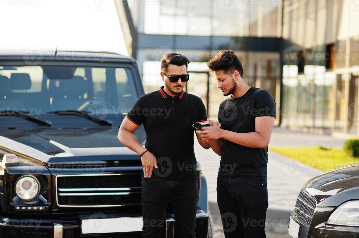Two asian brothers man wear on all black posed near suv cars and looking on mobile phone. photo