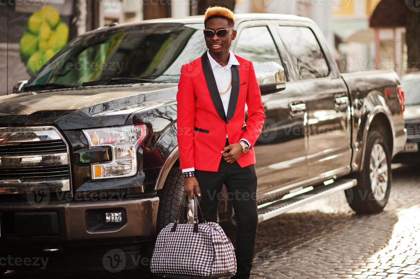 Fashion african american man model at red suit, with highlights hair posed against big suv black car. photo