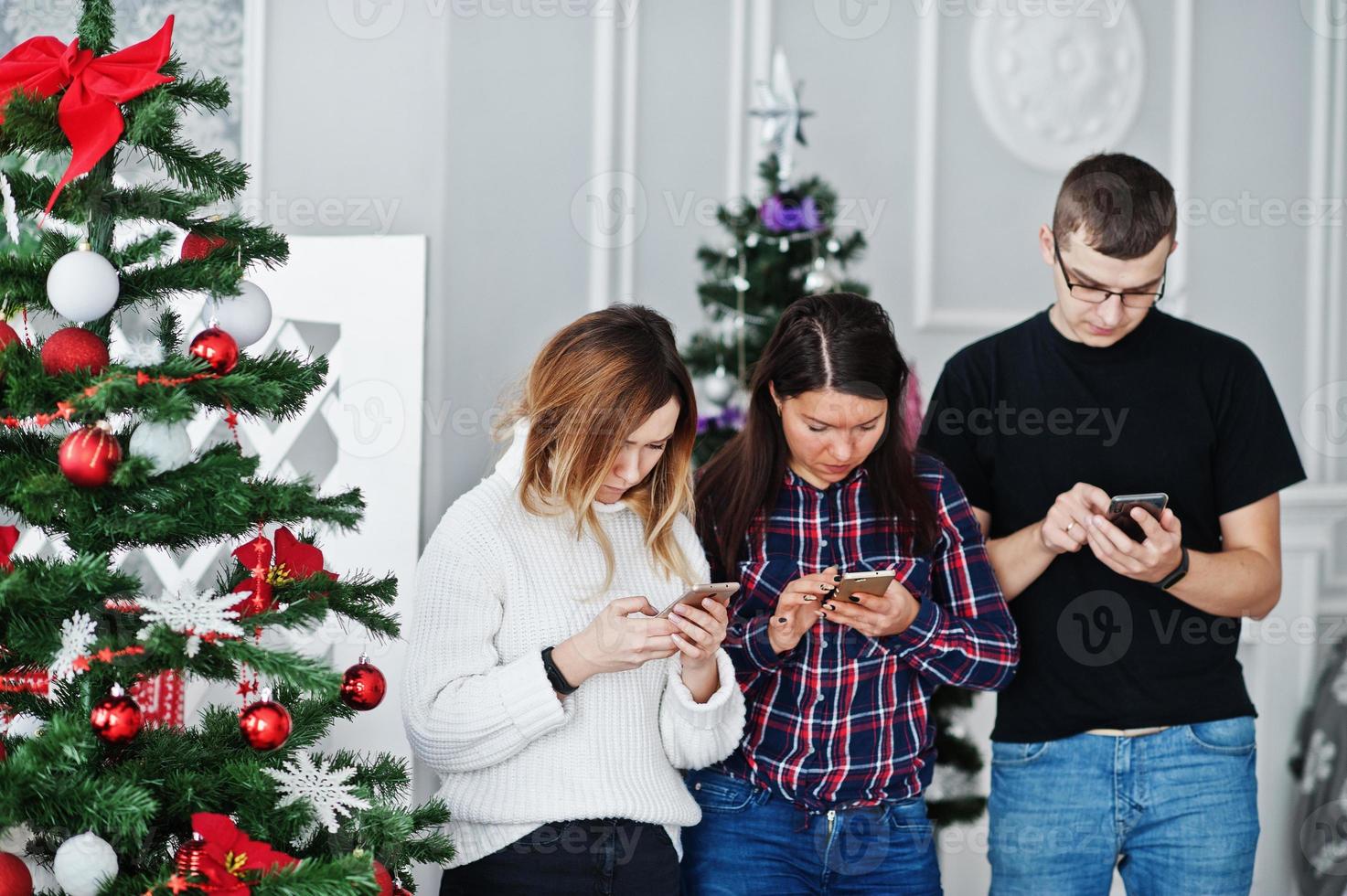 grupo de amigos como dos chicas y un hombre mirando teléfonos móviles contra el árbol de navidad en el estudio. foto