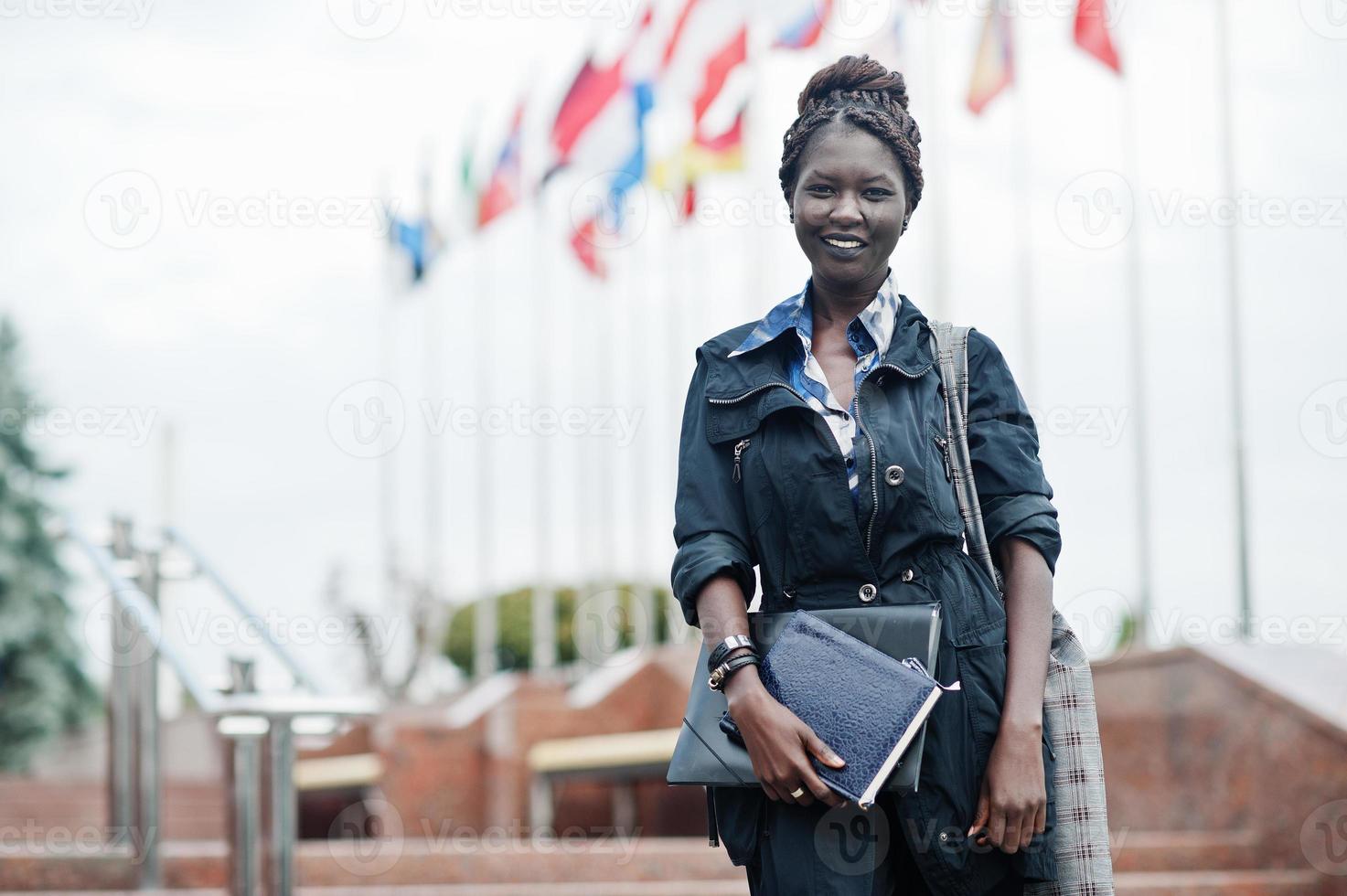 African student female posed with backpack and school items on yard of university, against flags of different countries. photo
