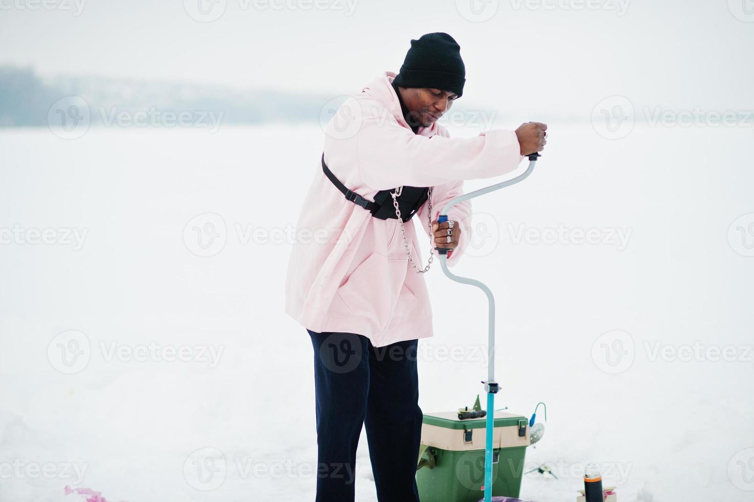 African american fisherman making hole in frozen ice by drill. Winter fishing. photo