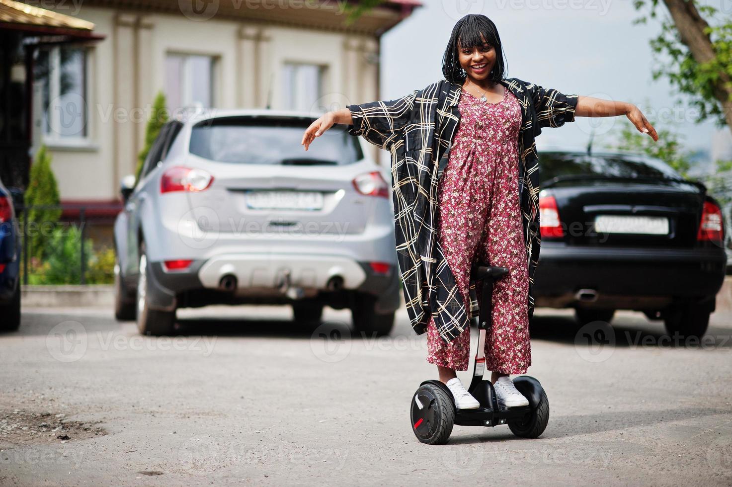 Beautiful african american woman using segway or hoverboard. Black girl on dual wheel self balancing electrical scooter against car parking. photo