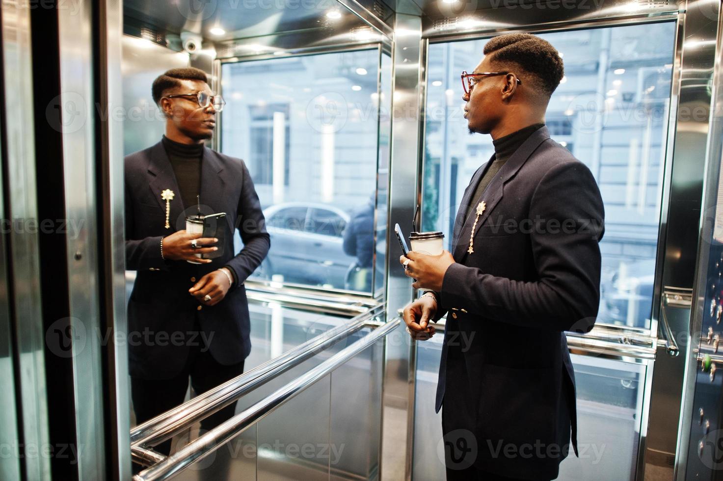 hombre afroamericano de moda con traje y gafas con teléfono móvil y una taza de café en las manos posado dentro del ascensor. foto