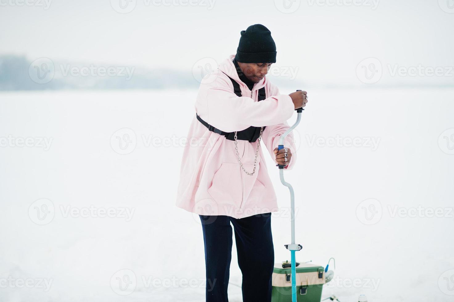 African american fisherman making hole in frozen ice by drill. Winter fishing. photo