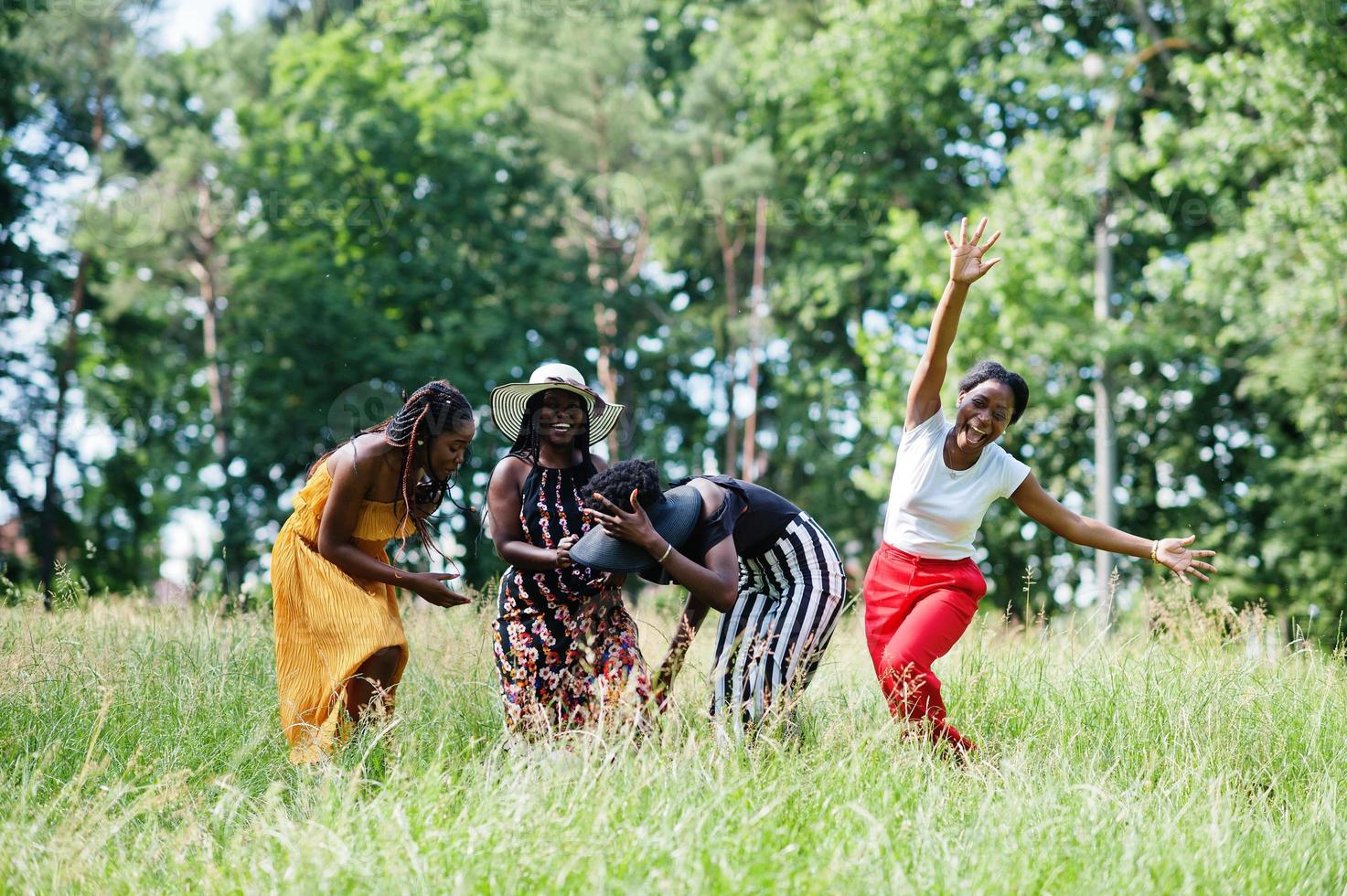 Group of four gorgeous african american womans wear summer hat jumping at green grass in park. photo