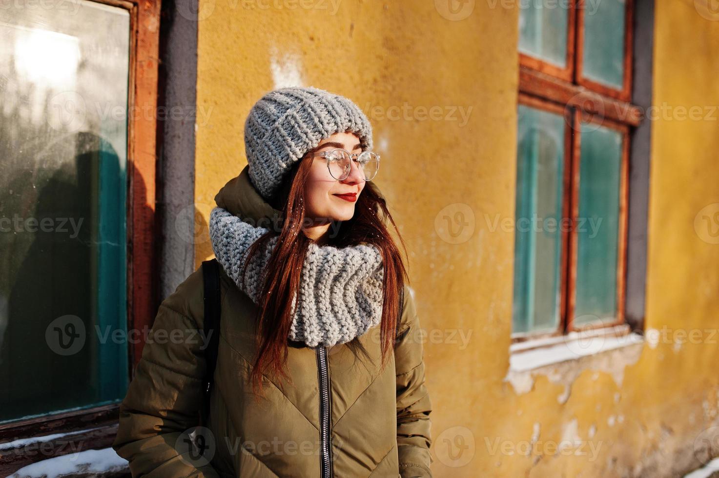 Portrait of brunette girl in gray scarf and hat, glasses at cold weather with sunshine against orange wall of old house. photo