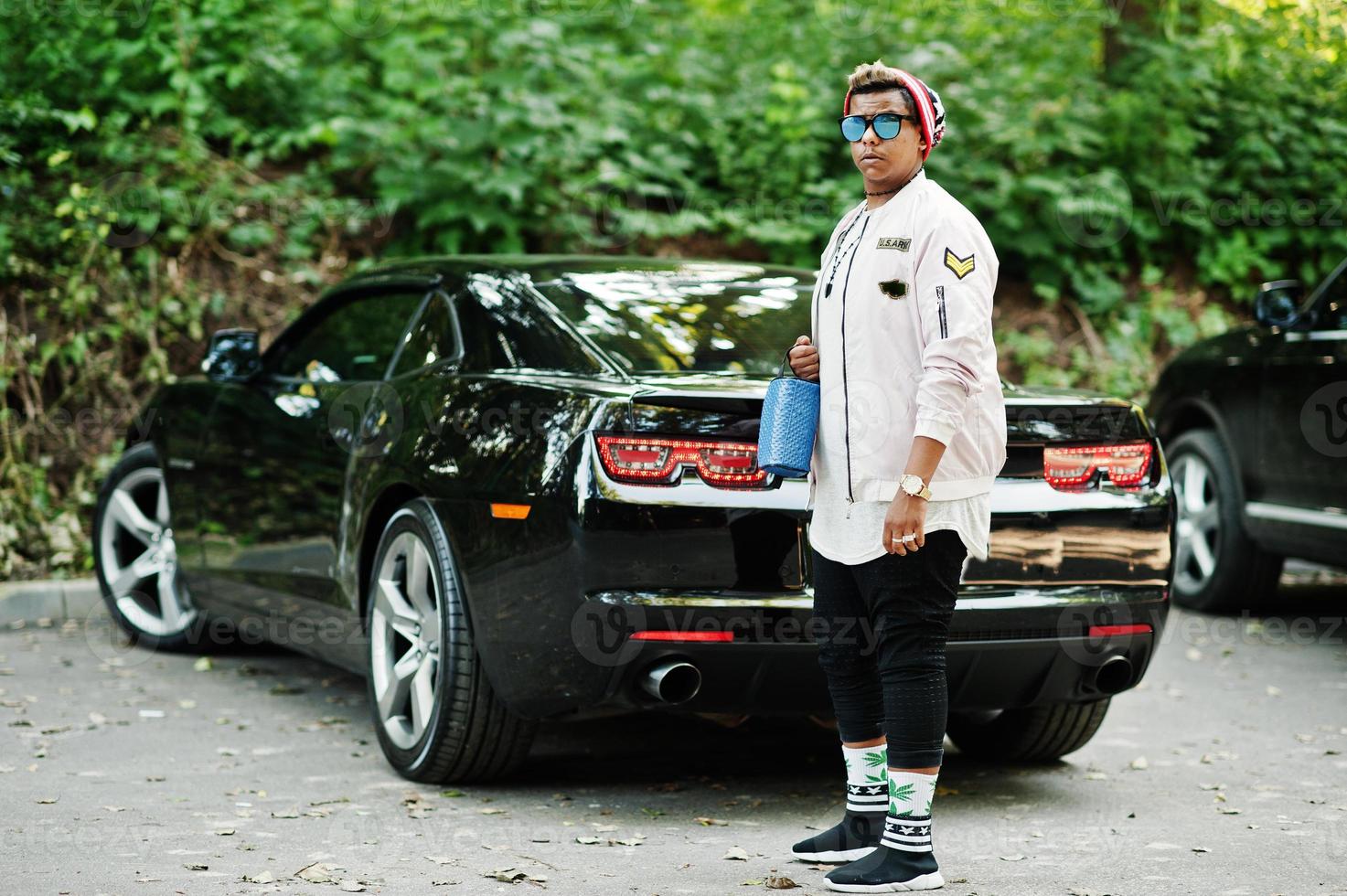 Stylish hipster arab man guy in hat and sunglasses posed outdoor in street against his black muscle car. photo