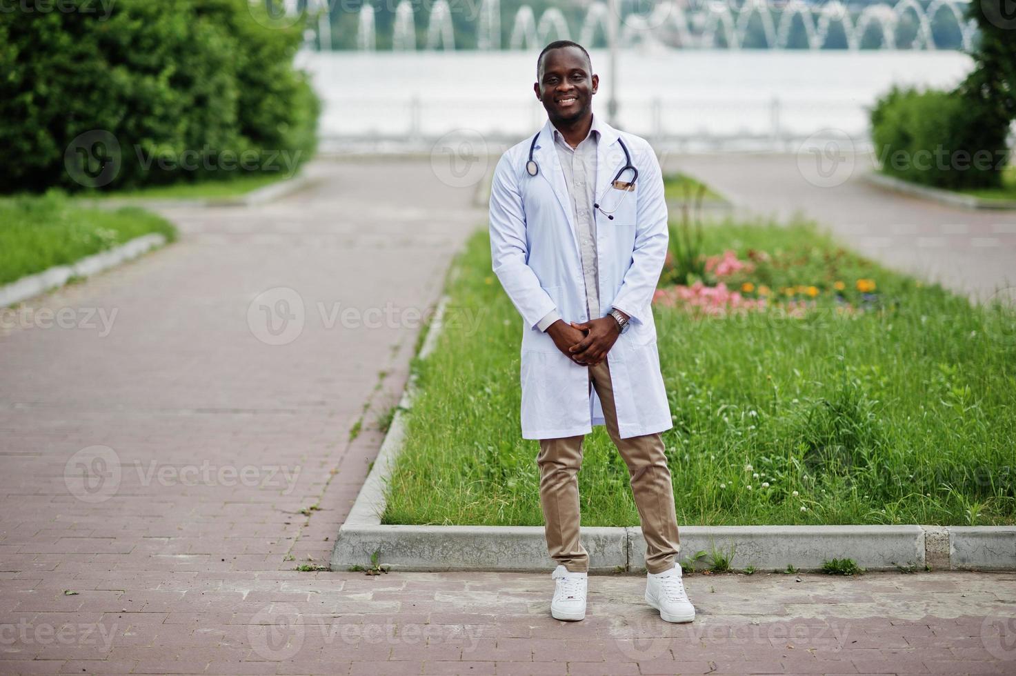 joven médico afroamericano de bata blanca con un estetoscopio posado al aire libre. foto