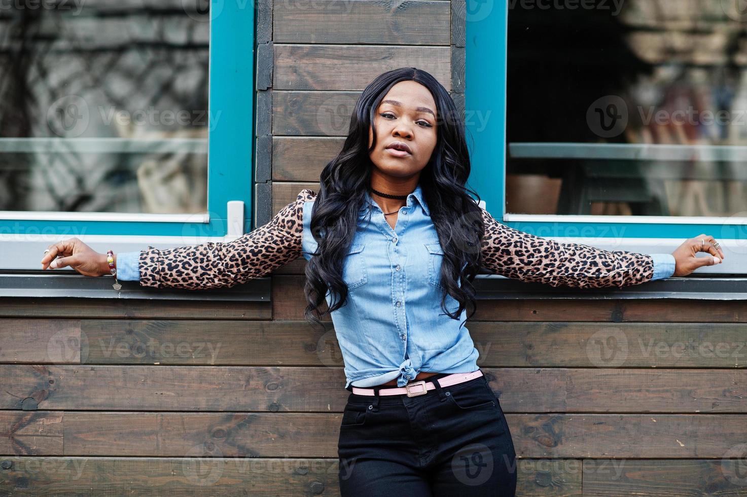 Hipster african american girl wearing jeans shirt with leopard sleeves posing at street against wooden house with windows. photo