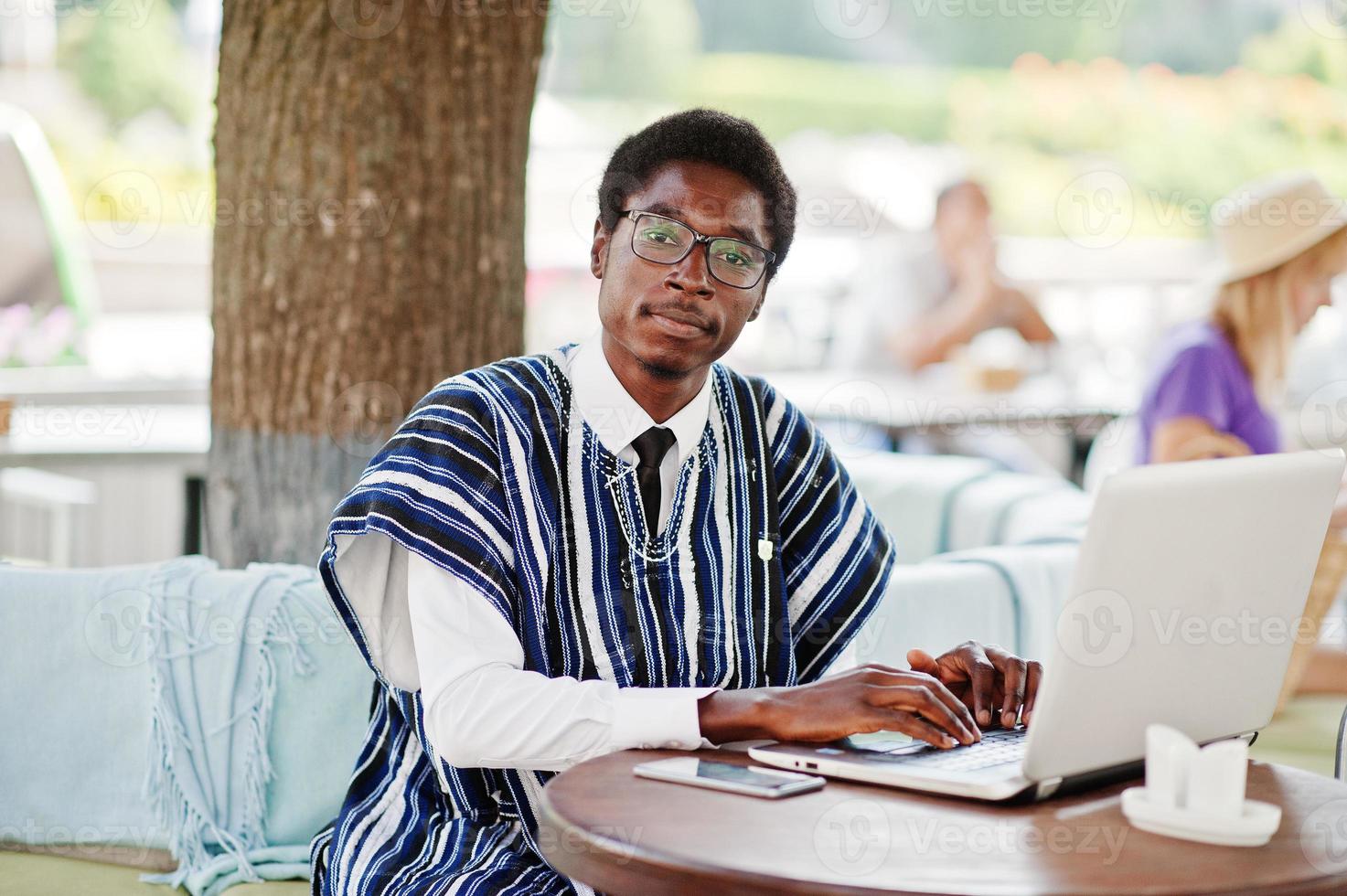 hombre africano con ropa tradicional y anteojos sentado detrás de una laptop en un café al aire libre y trabajando. foto