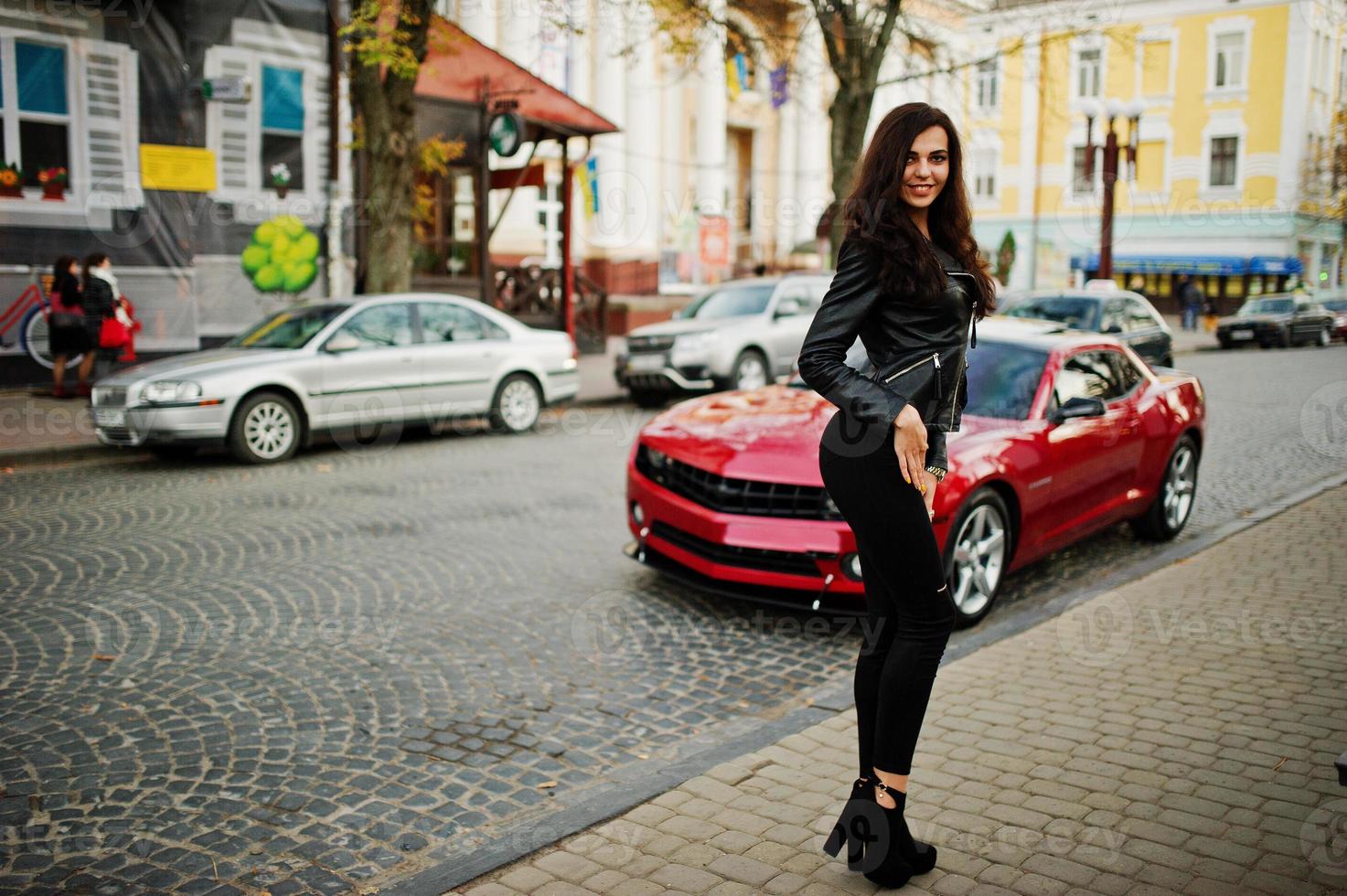 Young curly and sexy woman in leather jacket against red muscle car at street. photo
