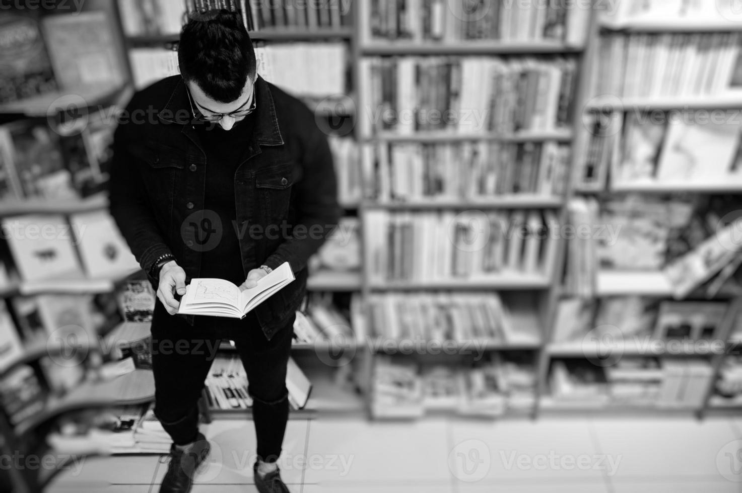 estudiante árabe alto e inteligente, vestido con chaqueta negra de jeans y anteojos, en la biblioteca con un libro en las manos. foto