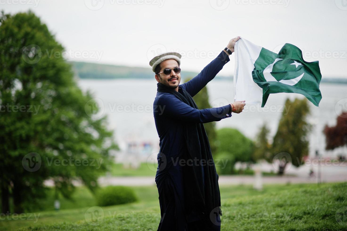 Stylish pakistani indian muslim arabic man in kurta dhoti suit, traditional pakol hat and sunglasses, hold Pakistan flag. photo