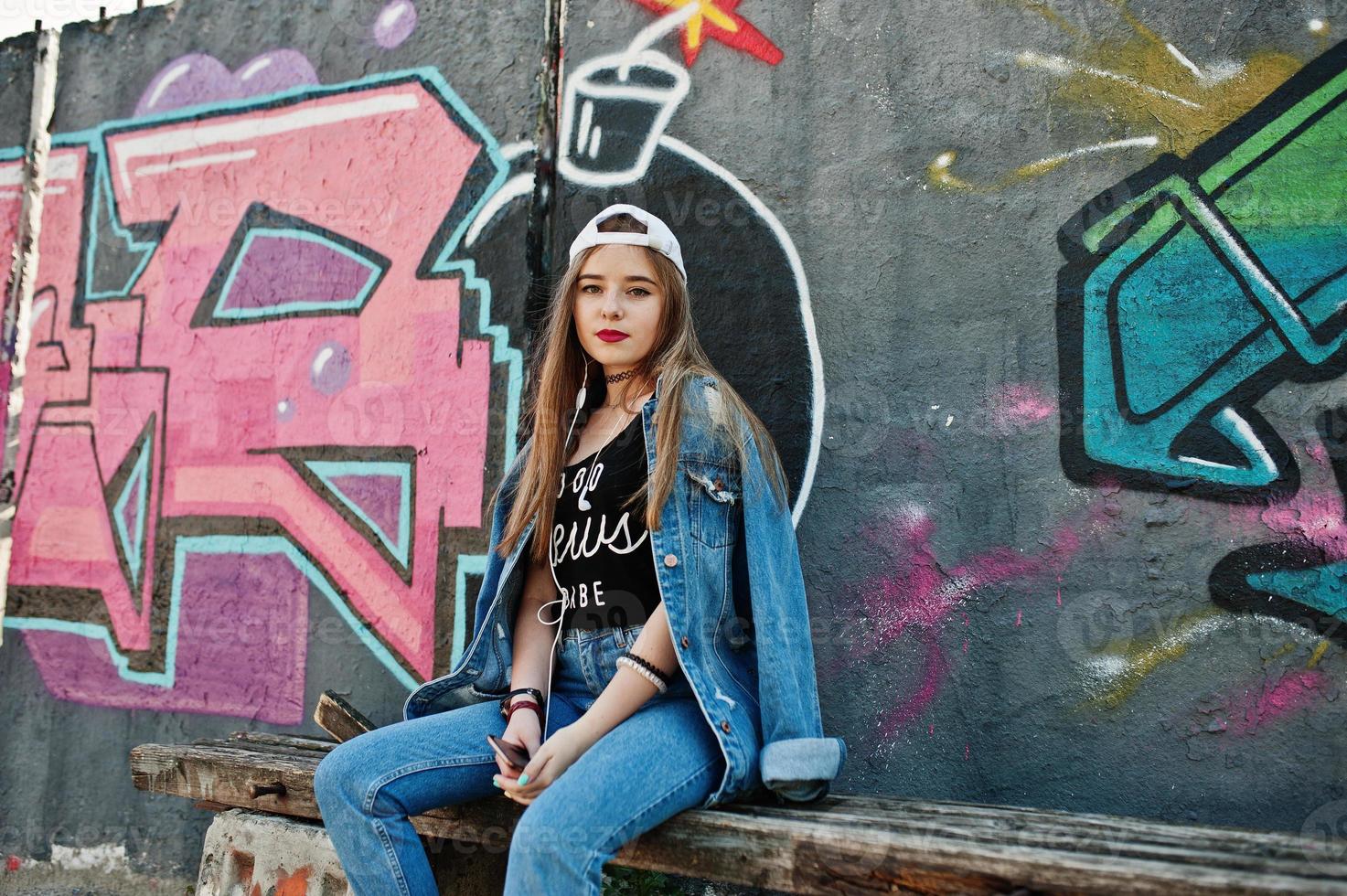 elegante chica hipster casual con gorra y jeans usa música para escuchar desde auriculares de teléfono móvil contra una gran pared de graffiti con bomba. foto