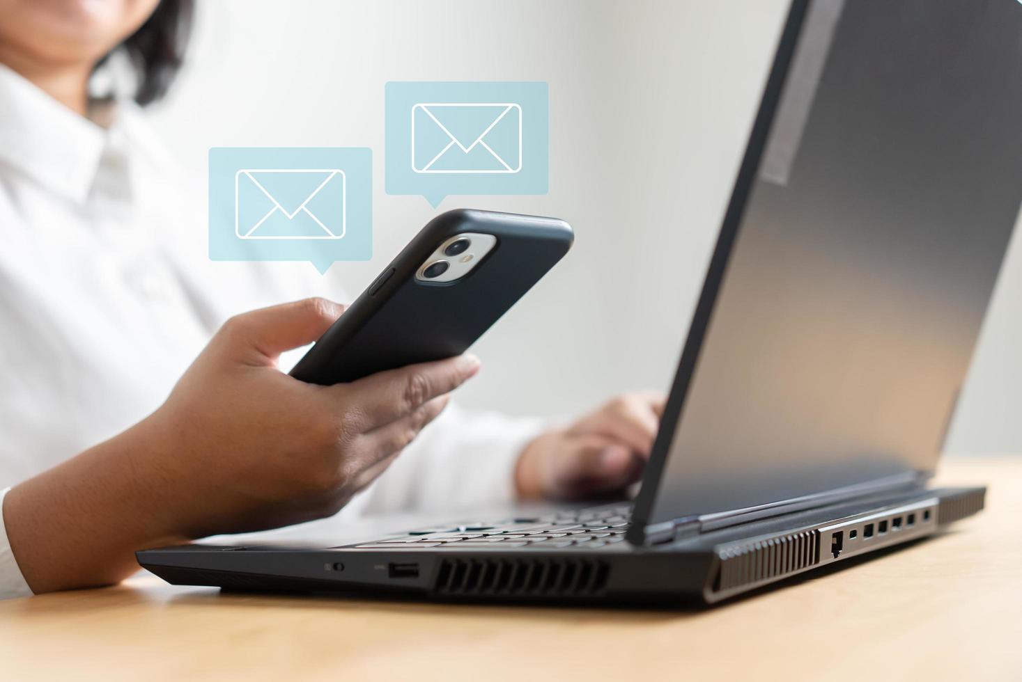 mano de mujer sosteniendo un teléfono móvil para recibir o enviar un icono de correo electrónico en el escritorio con una computadora portátil foto