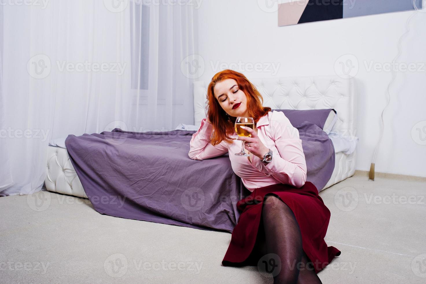 Gorgeous red haired girl in pink blouse and red skirt with glass of wine at hand near bed at room. photo