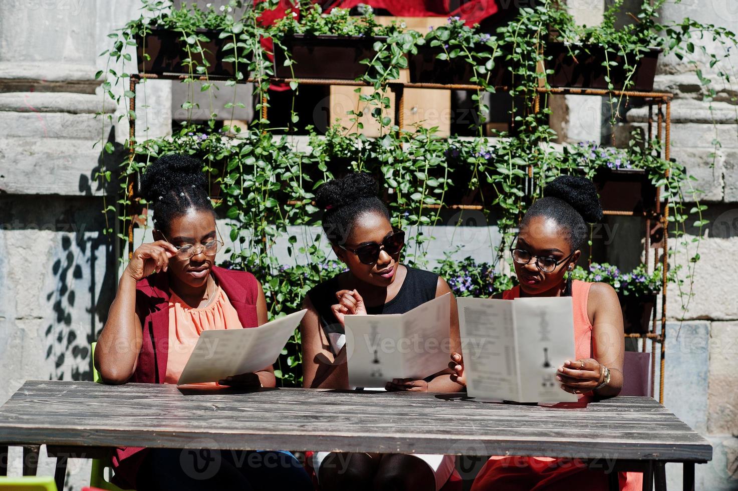 Three stylish african american womans posed at sunny summer day outdoor, sitting on table of restaurant with menu at hands. photo