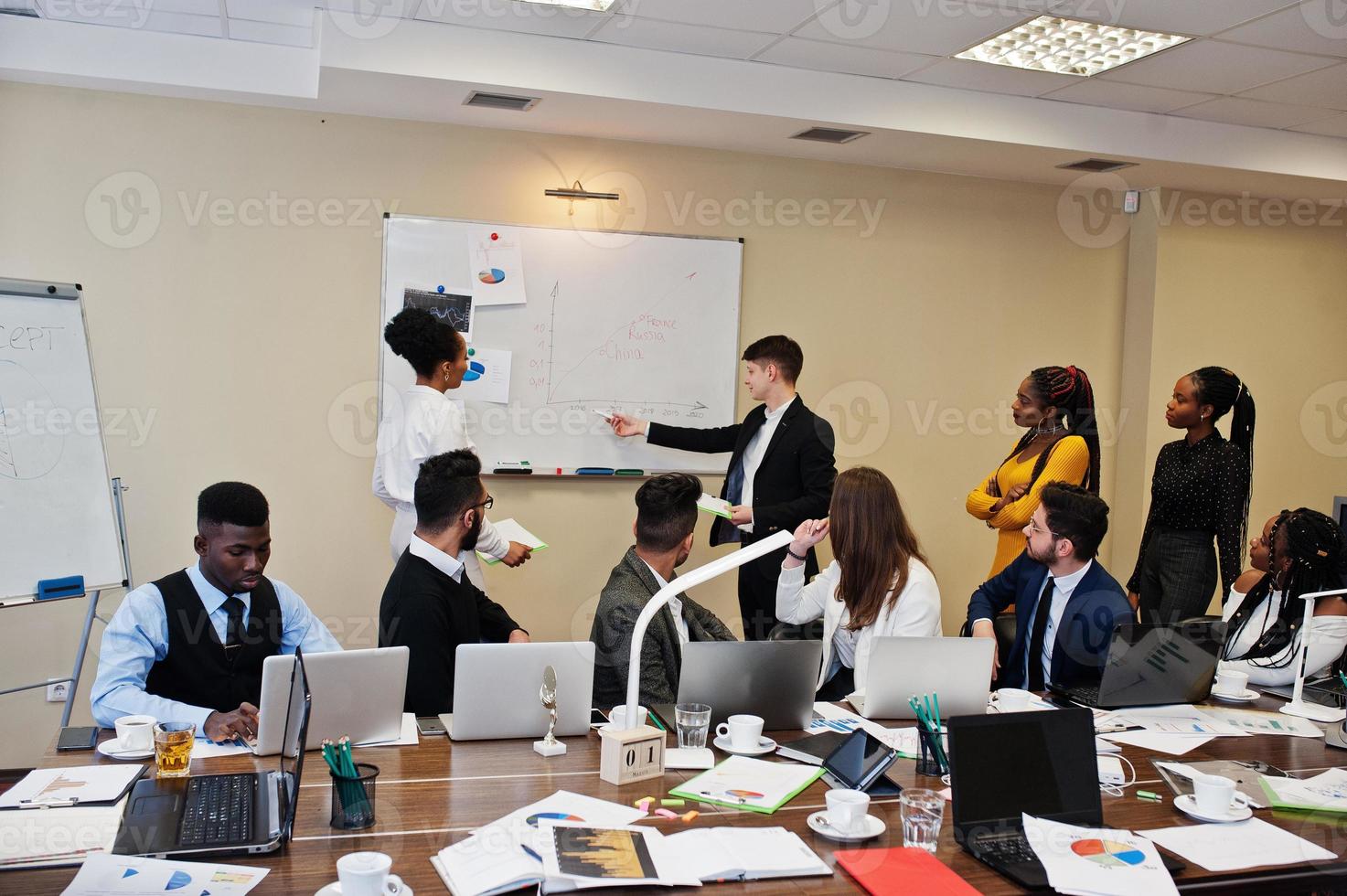 Mixed race business coach presenting report standing near whiteboard pointing on sales statistic shown on diagram and chart teach diverse company members gathered together in conference room. photo