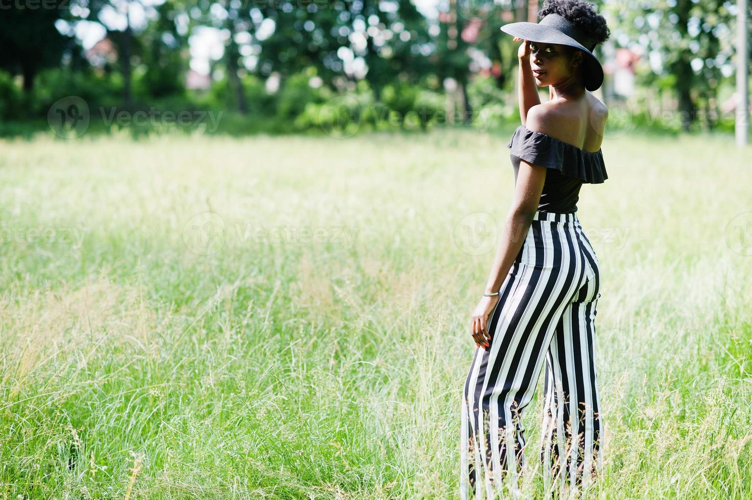 Portrait of gorgeous african american woman 20s in wear in black and white stripe pants and summer hat posing at green grass in park. photo