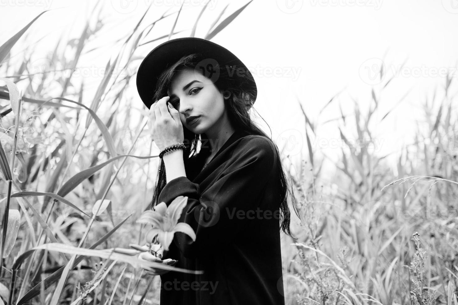 Sensual girl all in black, red lips and hat. Goth dramatic woman hold orange lily flower on  common reed. Black and white portrait. photo