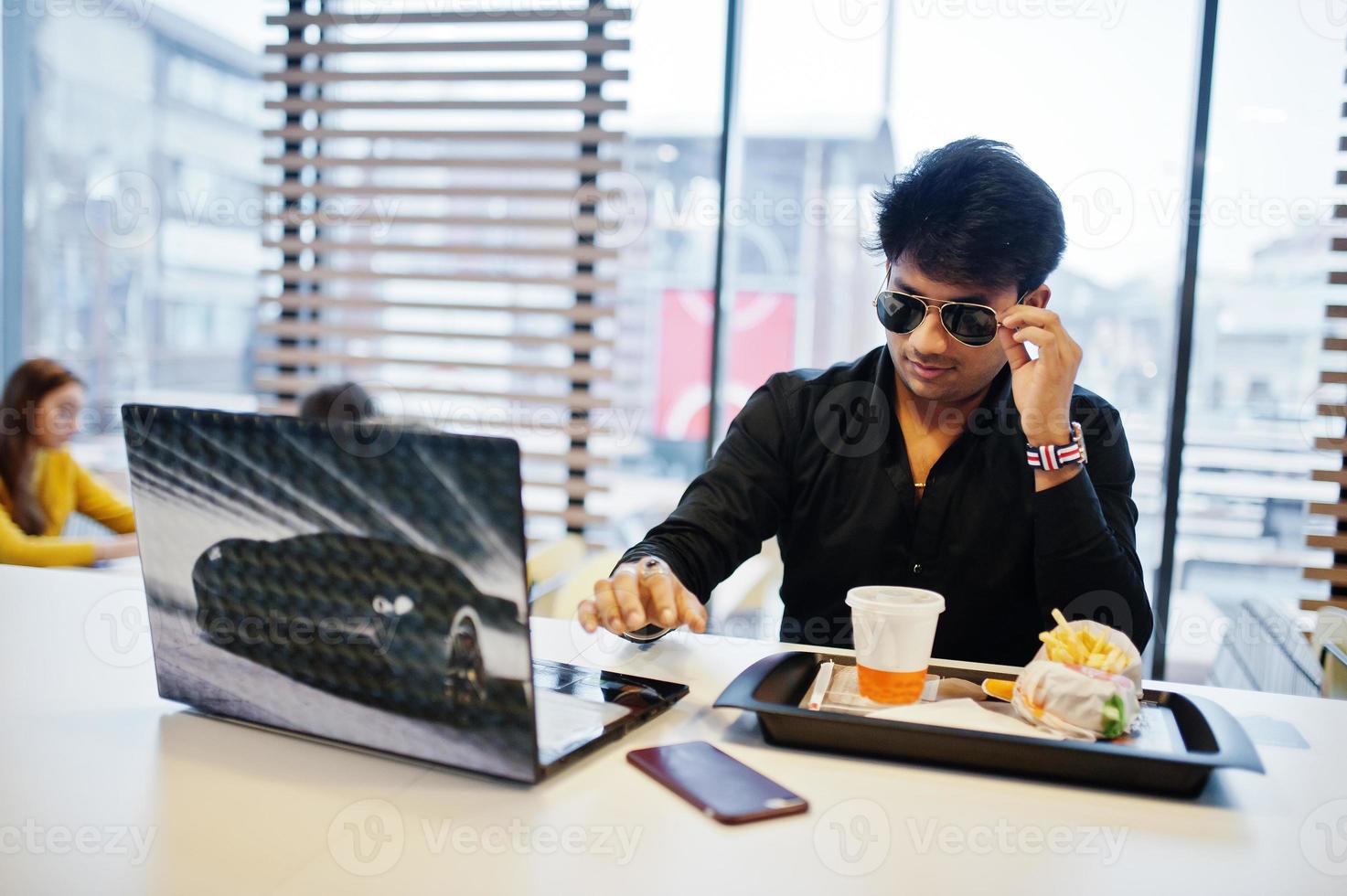 Stylish indian man in sunglasses sitting at fast food cafe against his laptop. photo