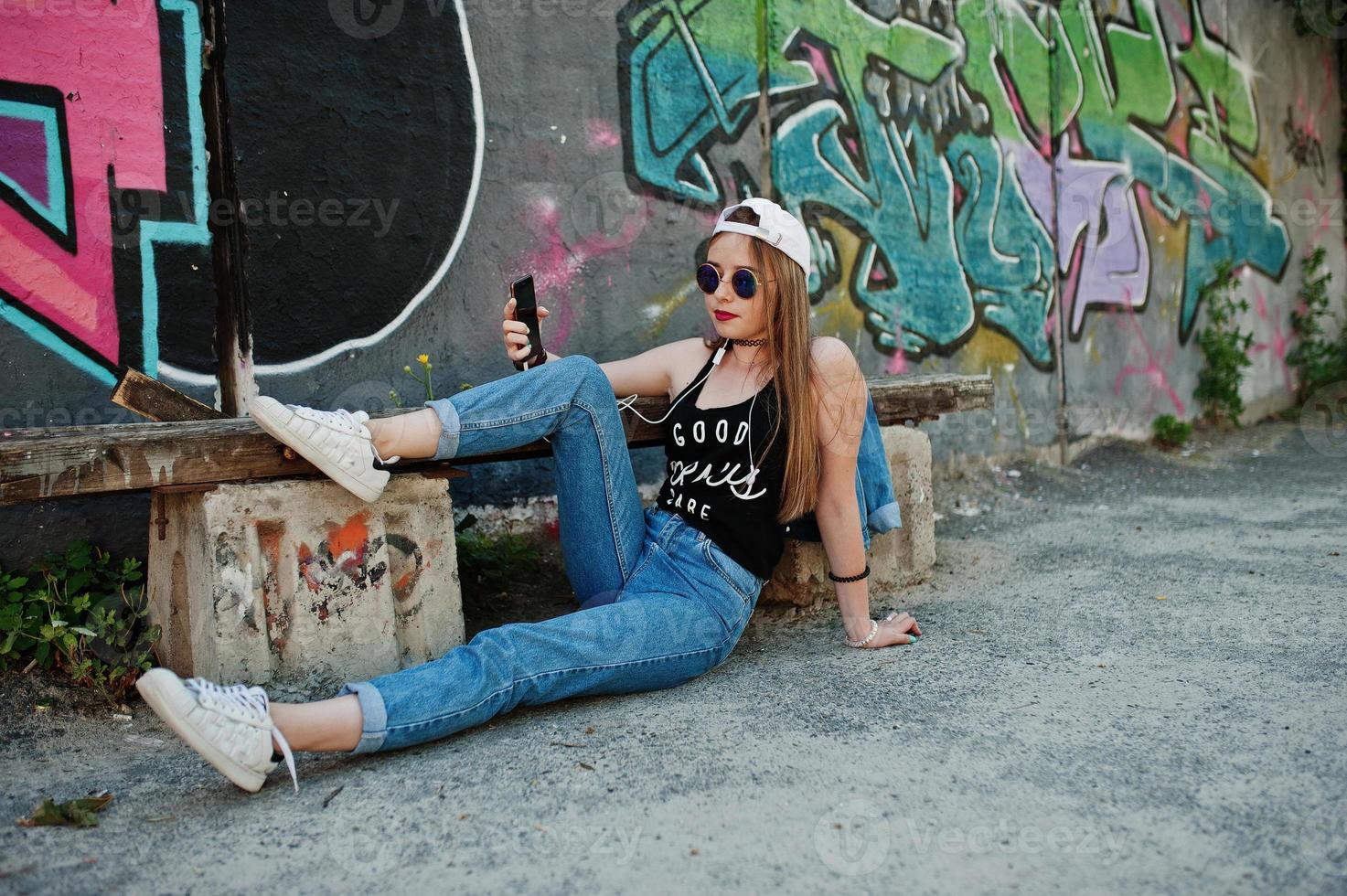 Stylish casual hipster girl in cap, sunglasses and jeans wear, listening music from headphones of mobile phone against large graffiti wall. photo