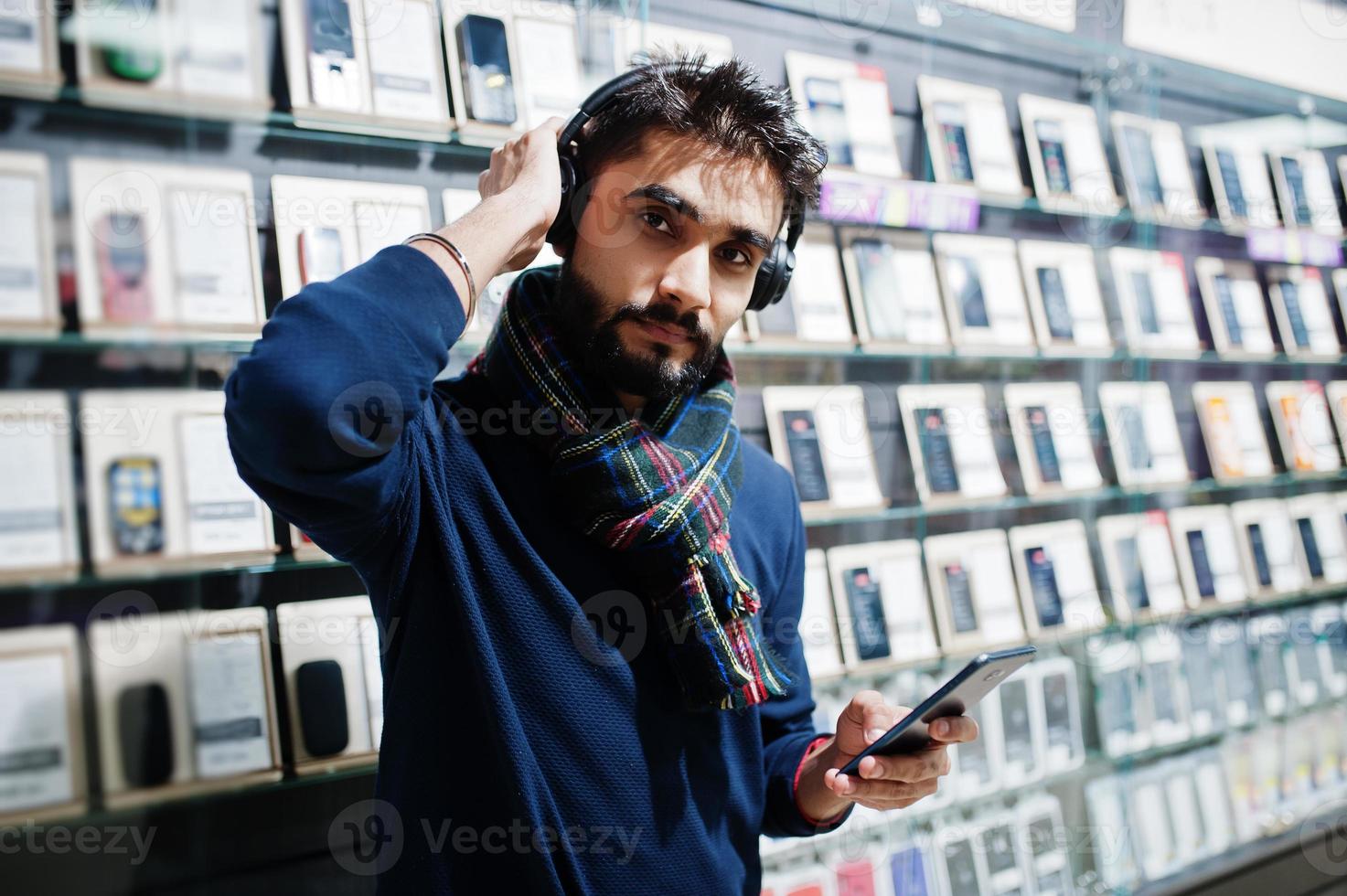 Indian beard man customer buyer at mobile phone store listen music from wireless earphones connect to his smartphone. South asian peoples and technologies concept. Cellphone shop. photo