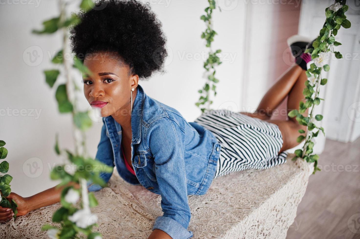 Attractive african american woman with afro hair wear on skirt and jeans jacket, posed at white room on swing. Fashionable black model. photo