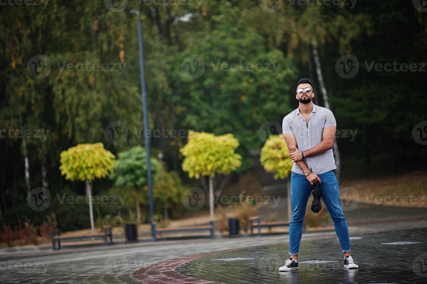 hombre de barba árabe alto de moda usa camisa, jeans y gafas de sol con paraguas posado bajo la lluvia en la plaza del parque. foto