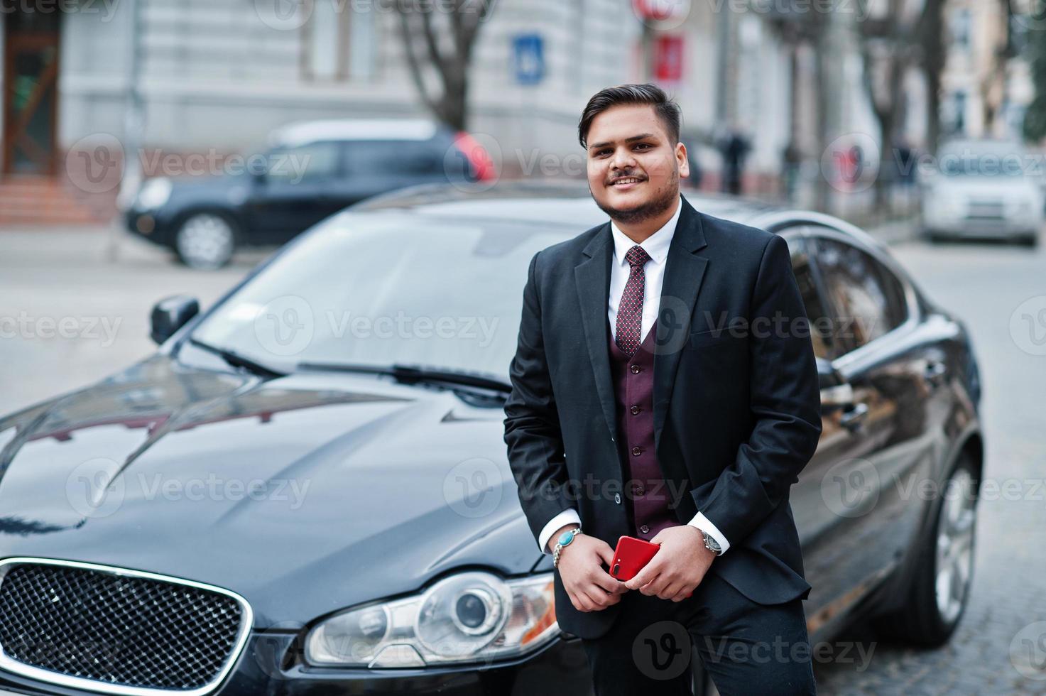 Stylish indian businessman in formal wear with mobile phone standing against black business car on street of city. photo