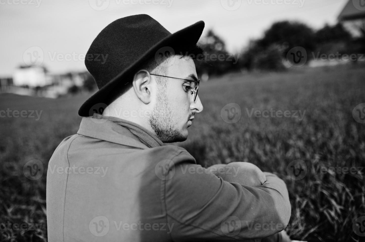 Stylish man in glasses, brown jacket and hat with bag posed on green field. photo