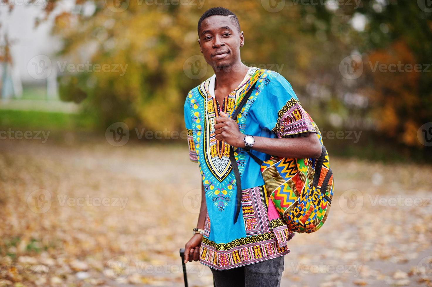 hombre africano en camisa tradicional africana en el parque de otoño con mochila y maleta. viajero emigrante. foto