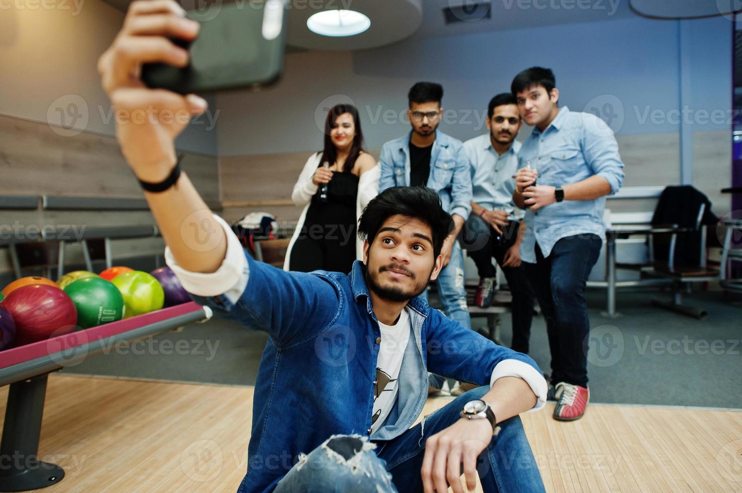 Group of five south asian peoples having rest and fun at bowling club. Making selfie by phone and holding cold soda drinks from glass bottles. photo
