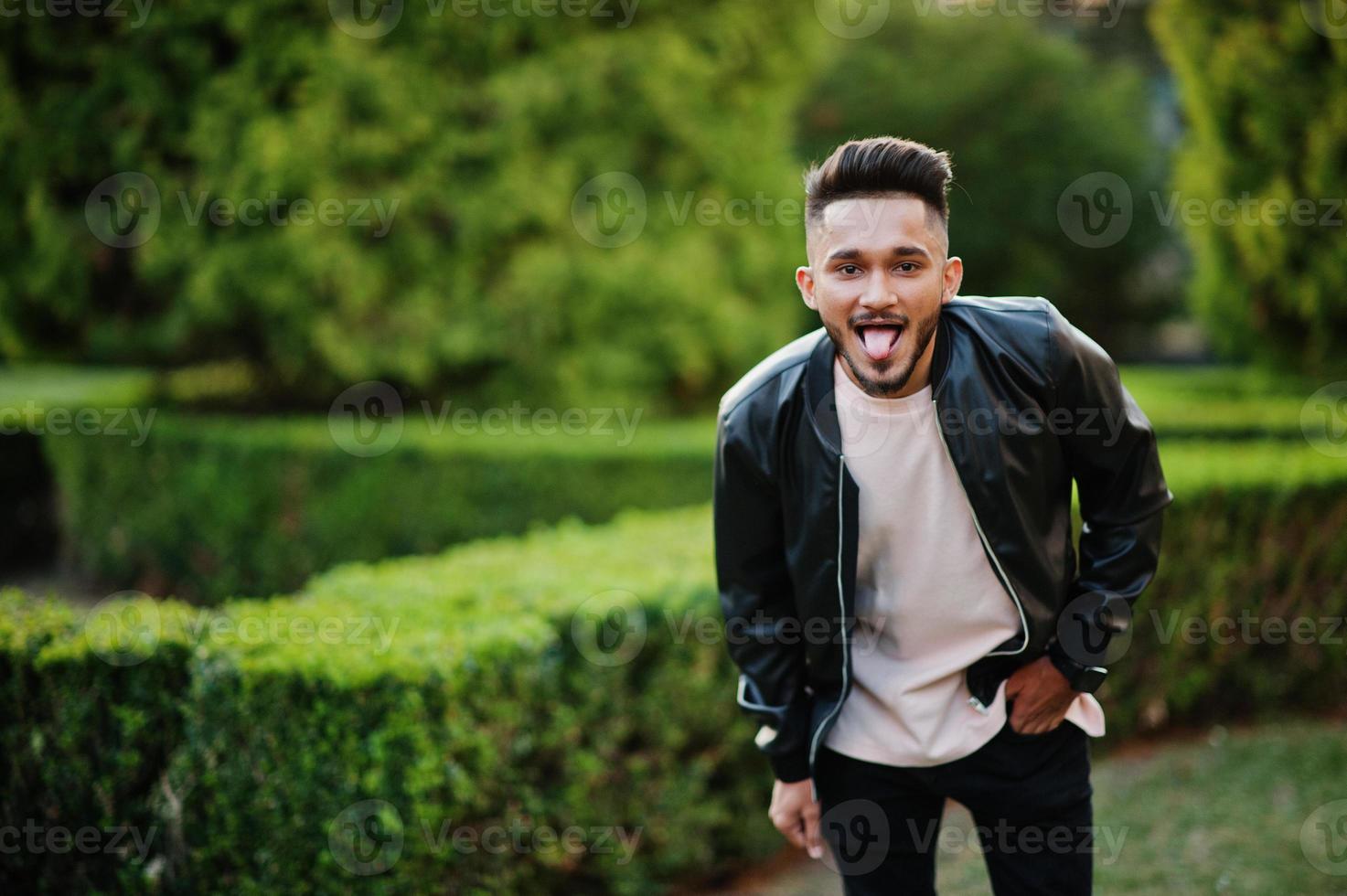 hombre con barba india con estilo en la chaqueta de cuero negro muestra tonque. modelo india posó en el jardín verde de la ciudad. foto