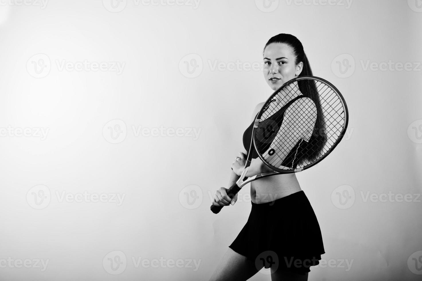 Black and white portrait of beautiful young woman player in sports clothes holding tennis racket while standing against white background. photo