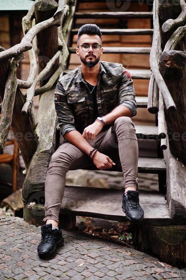 Impresionante hermoso hombre macho alto de barba árabe con gafas y chaqueta militar posado al aire libre contra escaleras de madera. foto