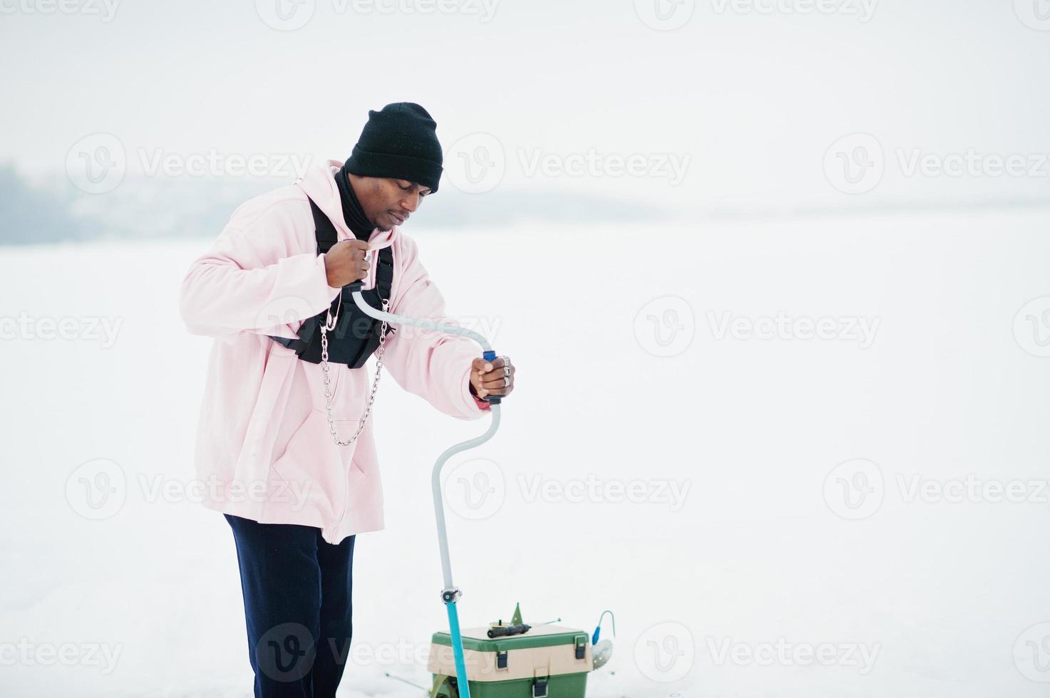 African american fisherman making hole in frozen ice by drill. Winter fishing. photo