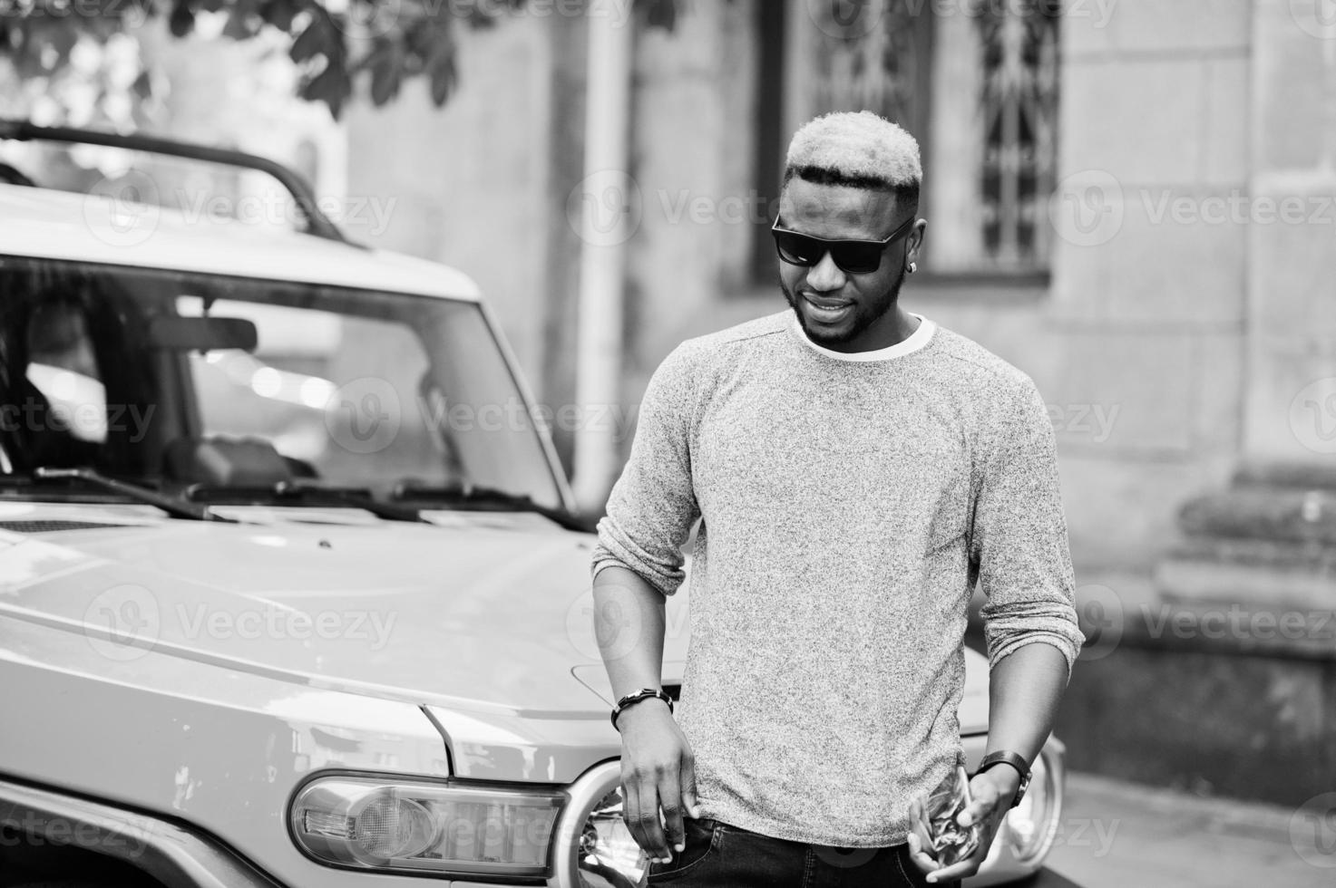 Stylish african american boy on gray sweater and black sunglasses posed on street against yellow car. Fashionable black business man. photo