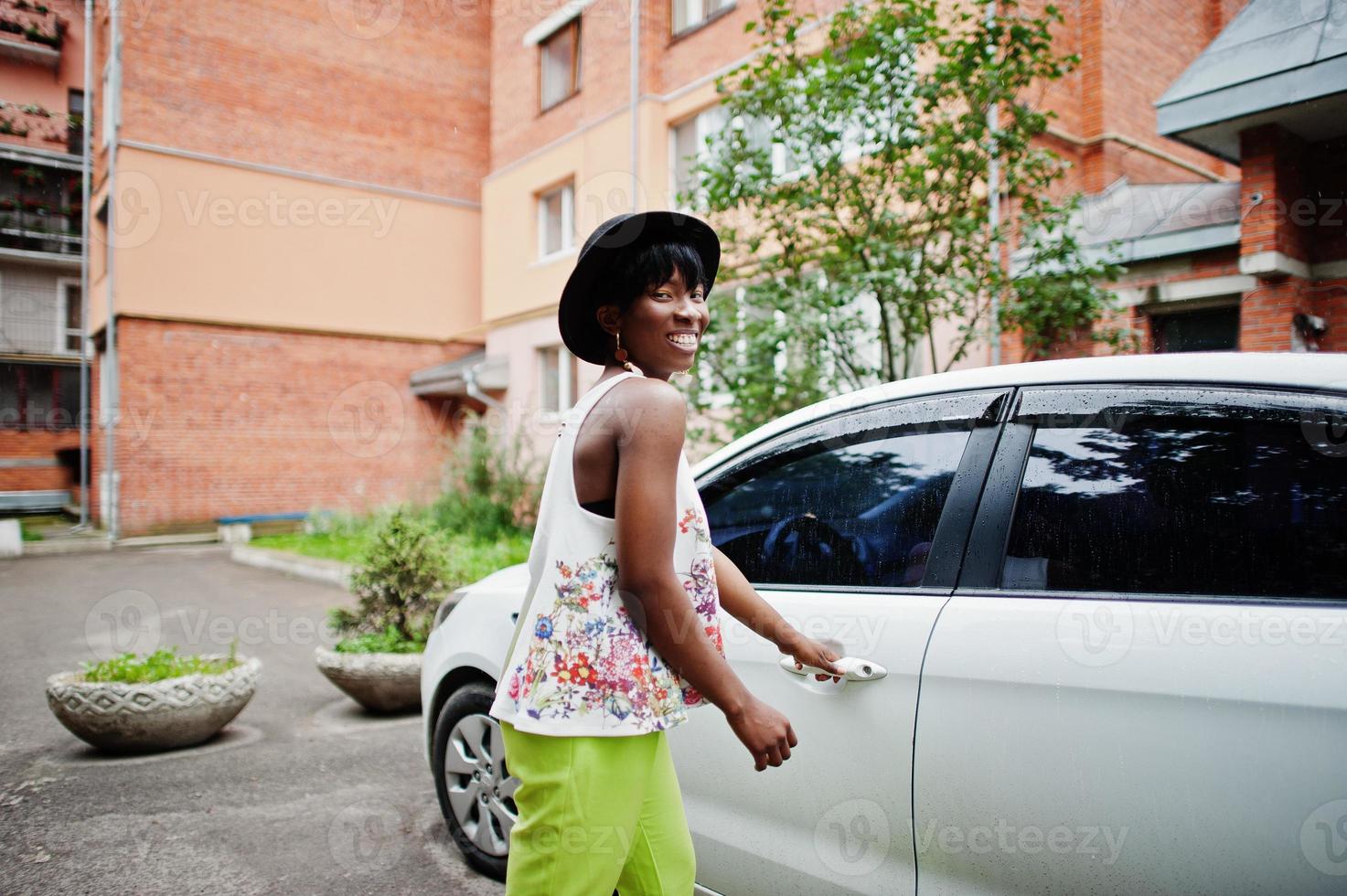 Amazing african american model woman in green pants and black hat posed near white door of car. photo