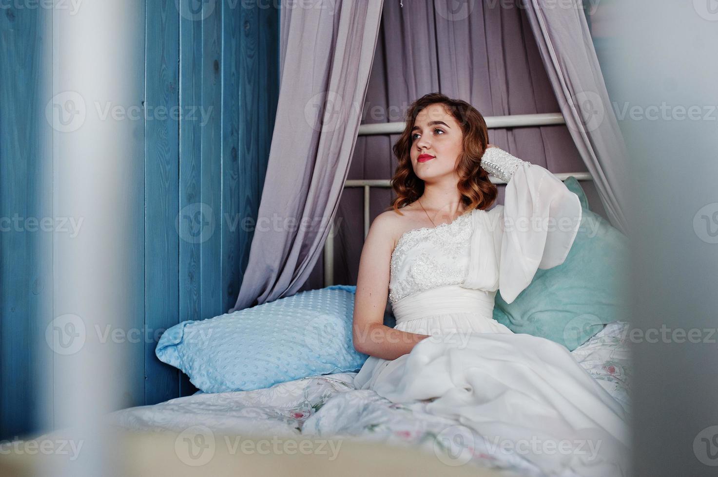 Pretty young curly gilrl in white dress sitting on bed. photo