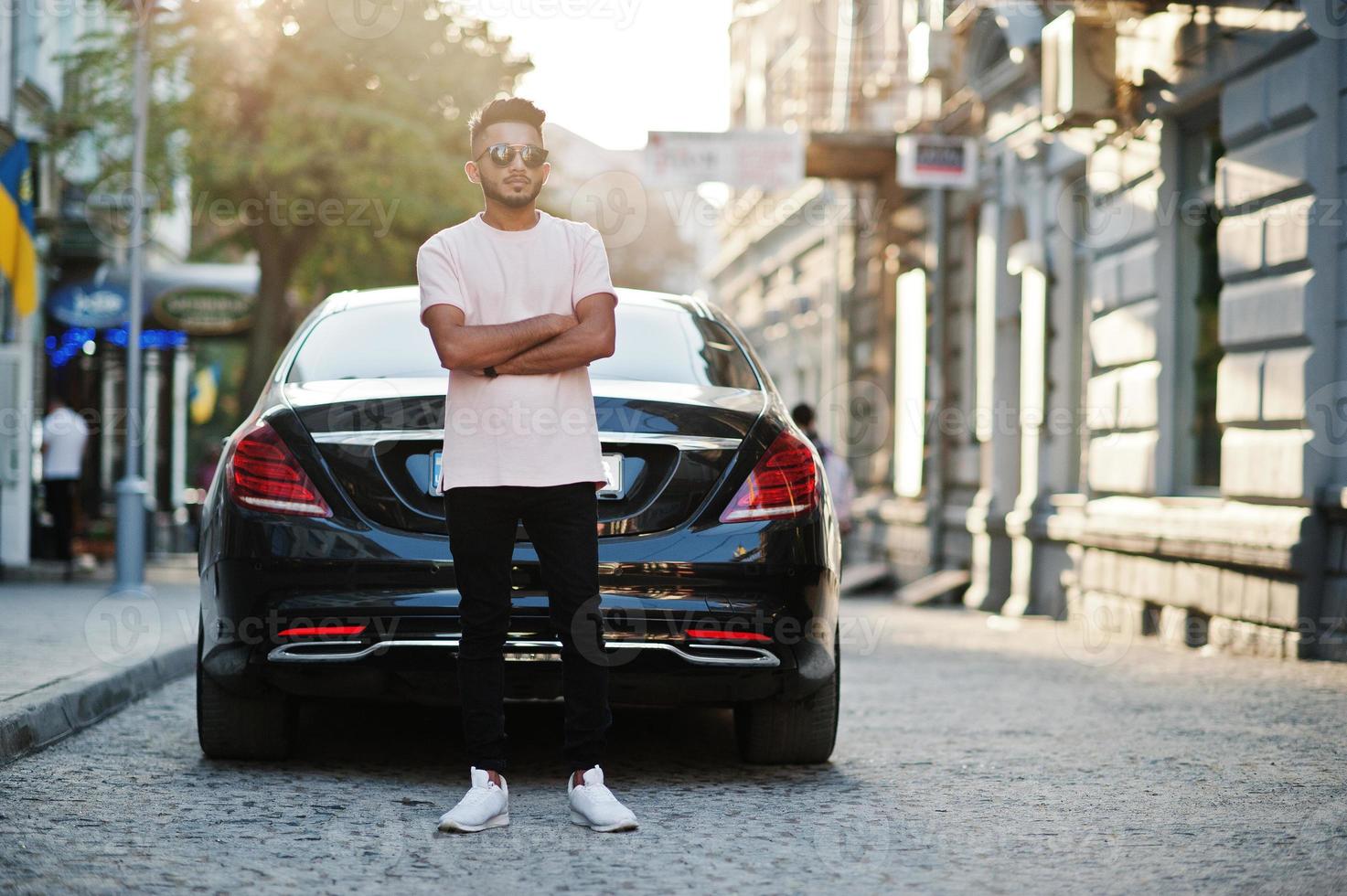 Stylish indian beard man at sunglasses and pink t-shirt against luxury car. India rich model posed outdoor at streets of city. photo