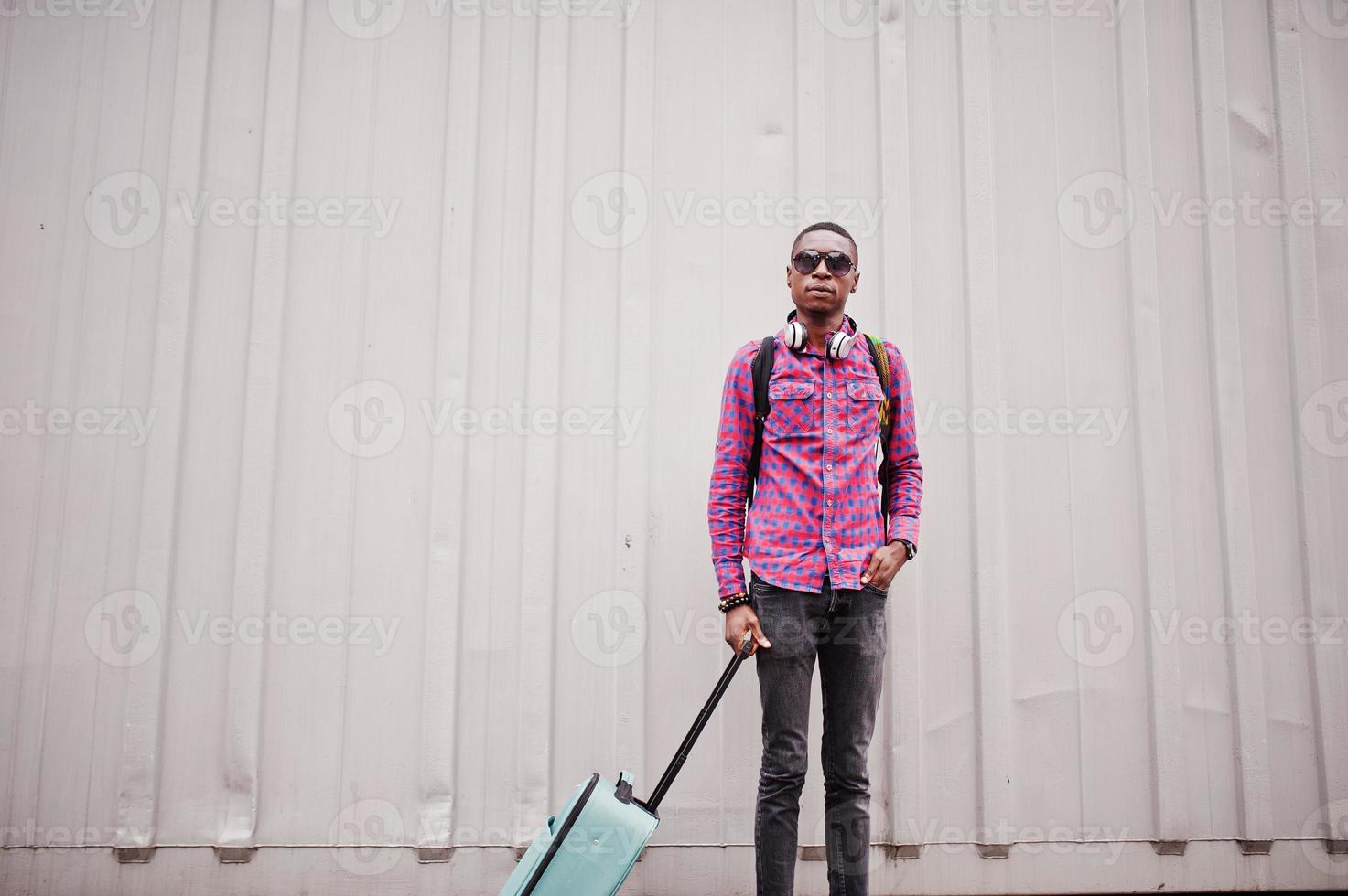 African american man in checkered shirt, sunglasses and earphones with suitcase and backpack. Black man traveler against wall. photo