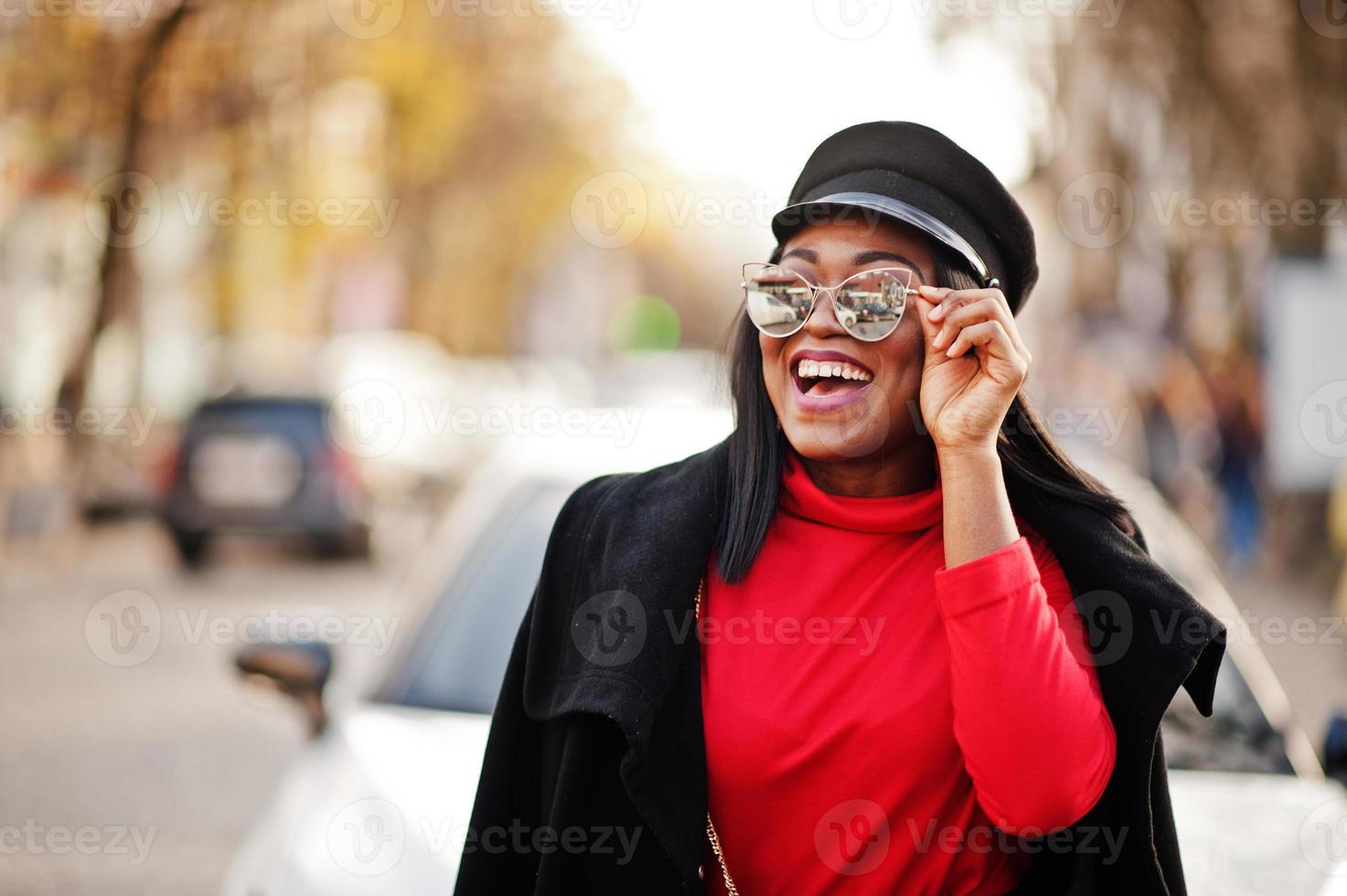 chica de moda afroamericana con abrigo, gorra de vendedor de periódicos y gafas de sol posadas en la calle contra un auto de negocios blanco. foto