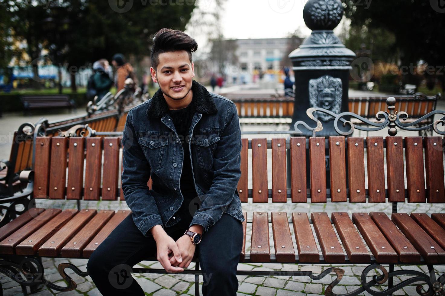 hombre indio guapo y de moda en chaqueta de jeans negros posó al aire libre, sentado en un banco. foto