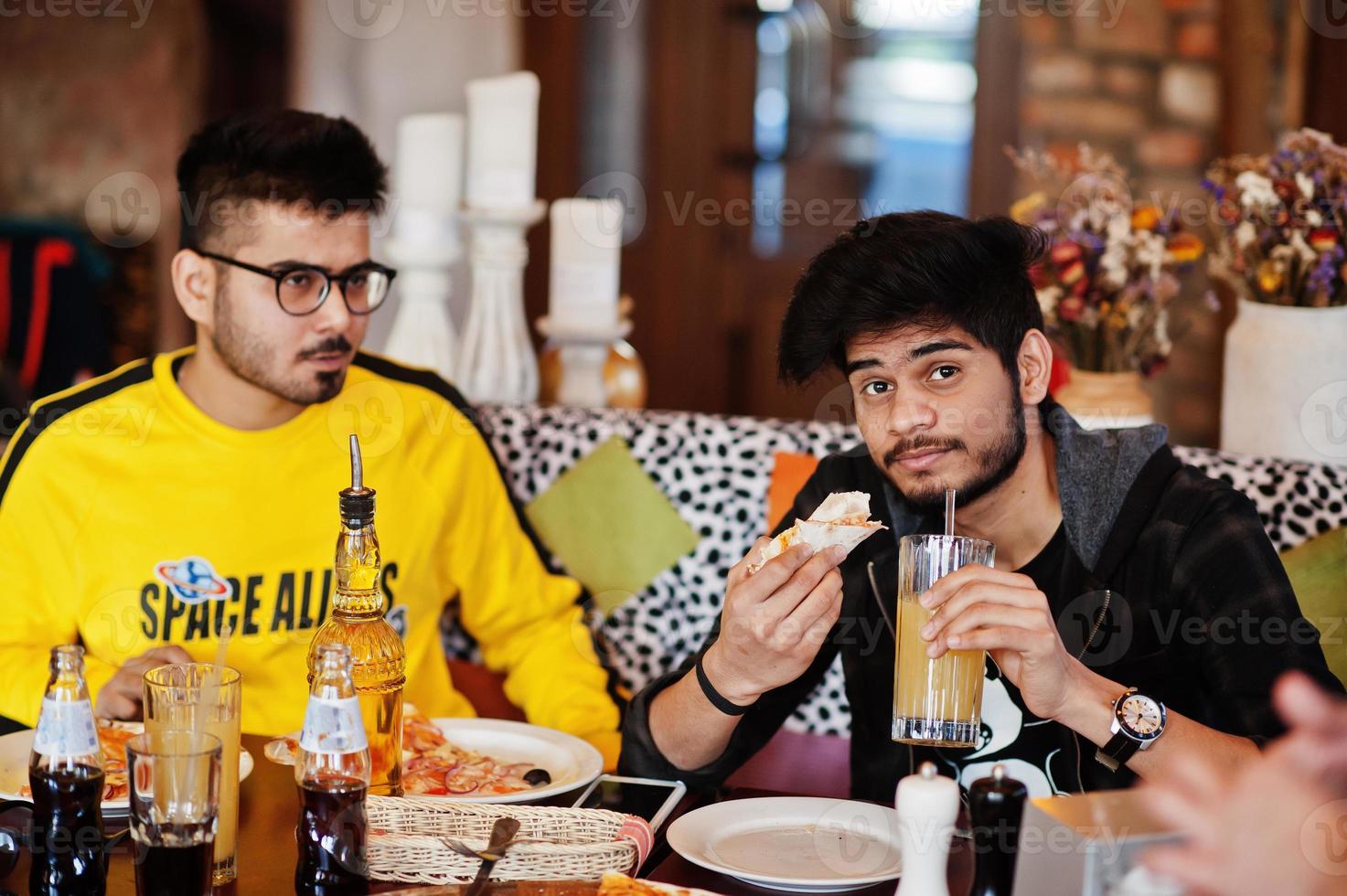 Asian friends guys eating pizza during party at pizzeria. Happy indian people having fun together, eating italian food and sitting on couch. photo