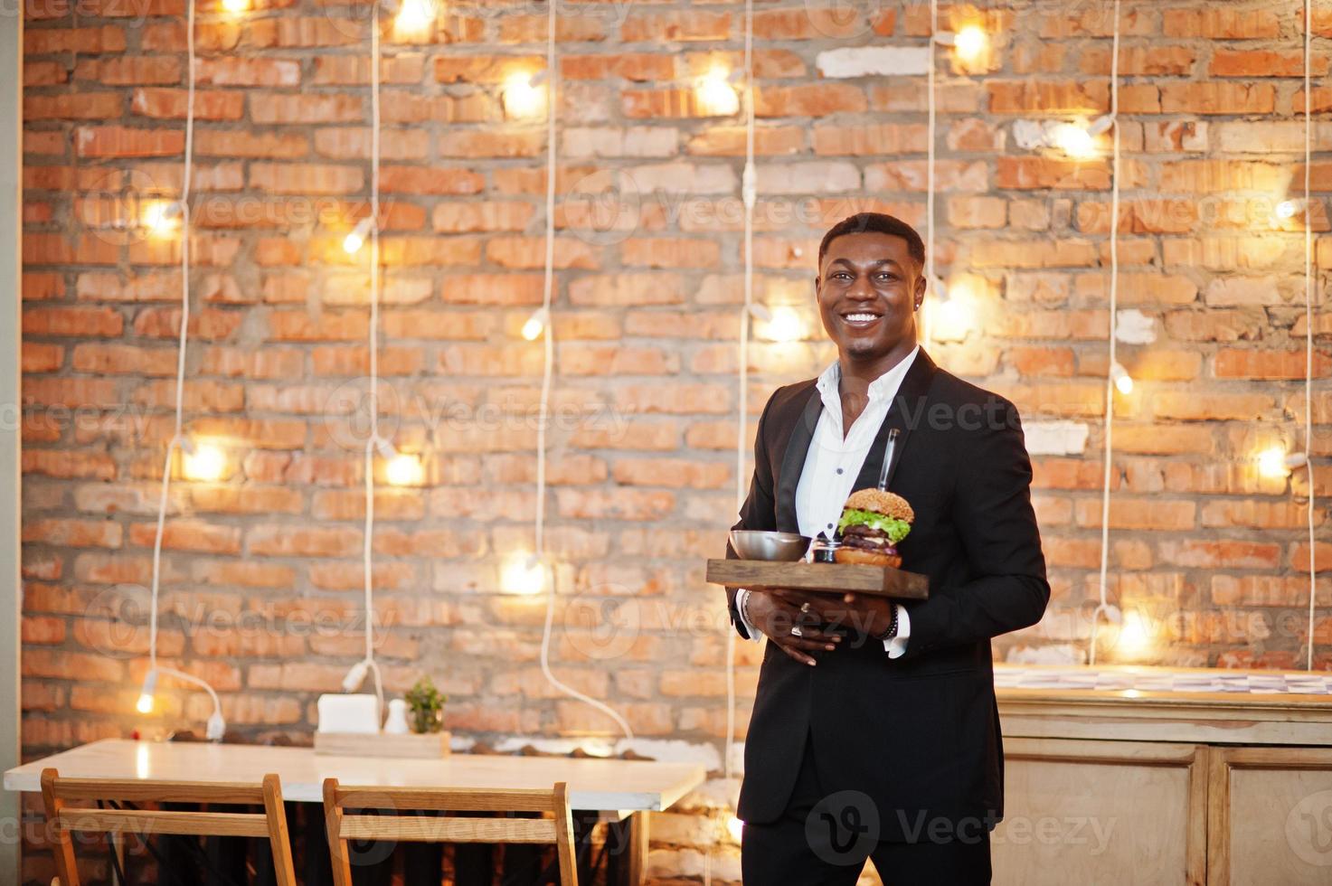 Respectable young african american man in black suit hold tray with double burger against brick wall of restaurant with lights. photo