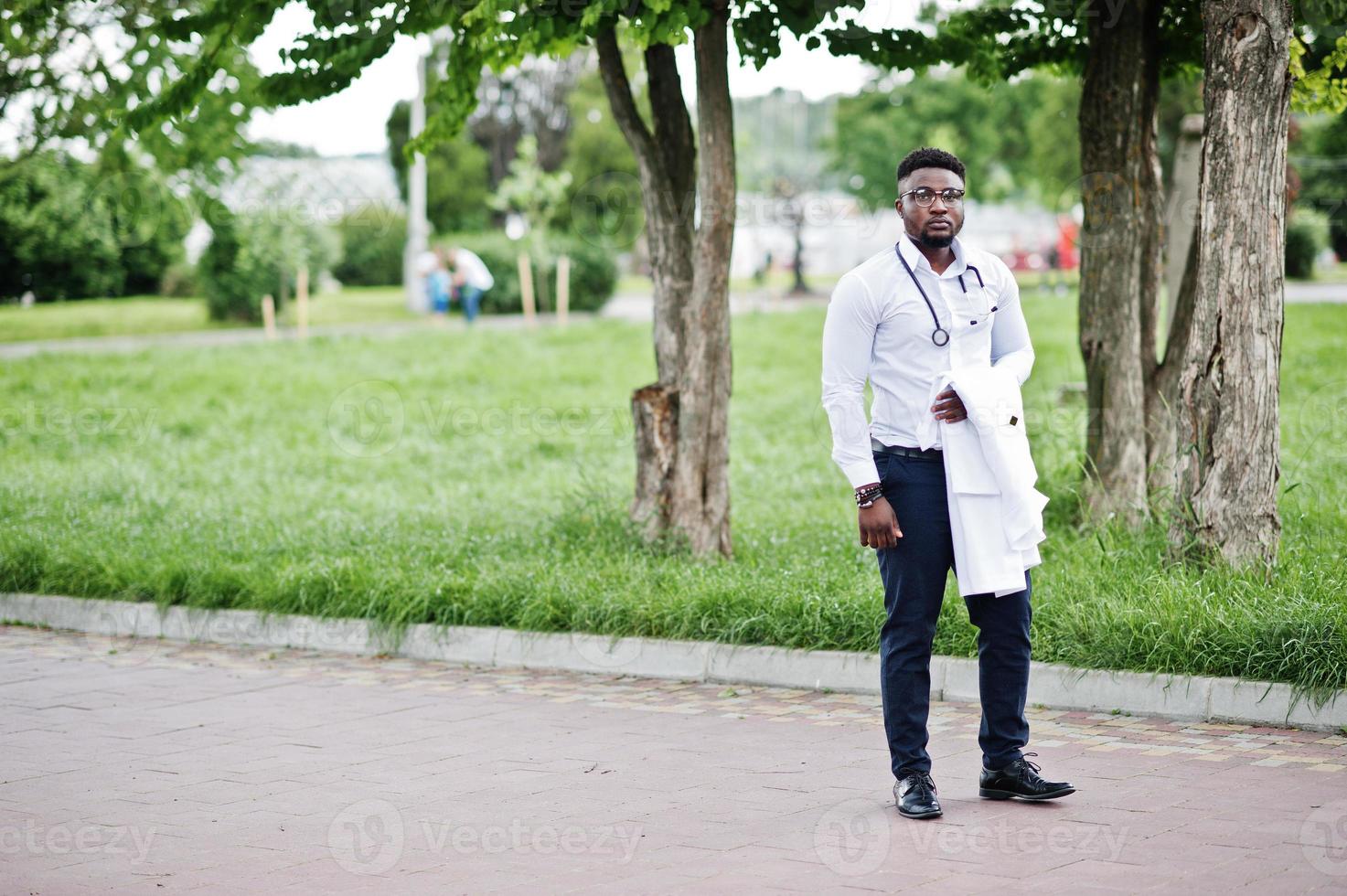un joven médico afroamericano sostiene una bata blanca a mano con un estetoscopio posado al aire libre. foto