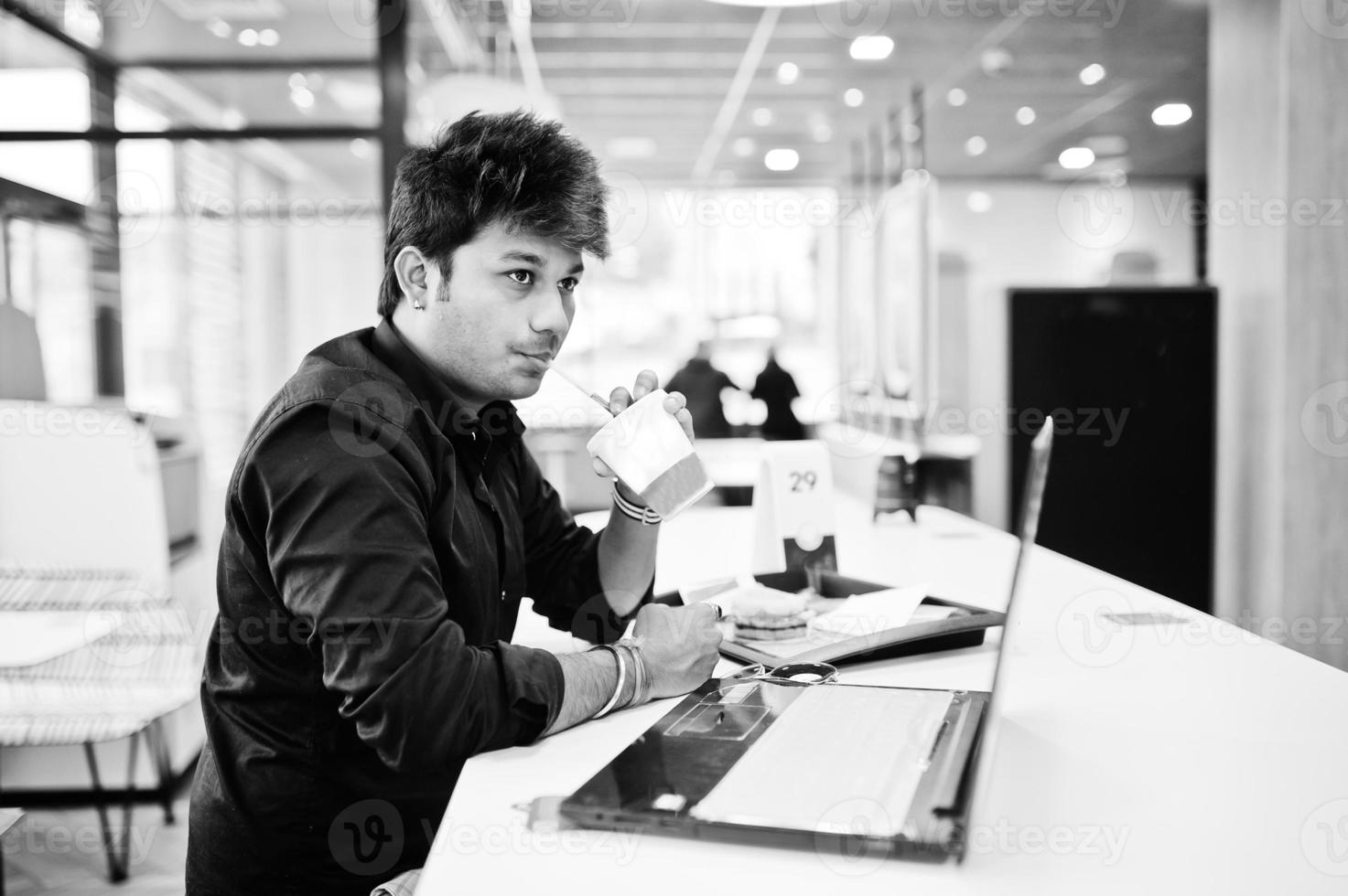 Stylish indian man sitting at fast food cafe against his laptop and drink soda. photo