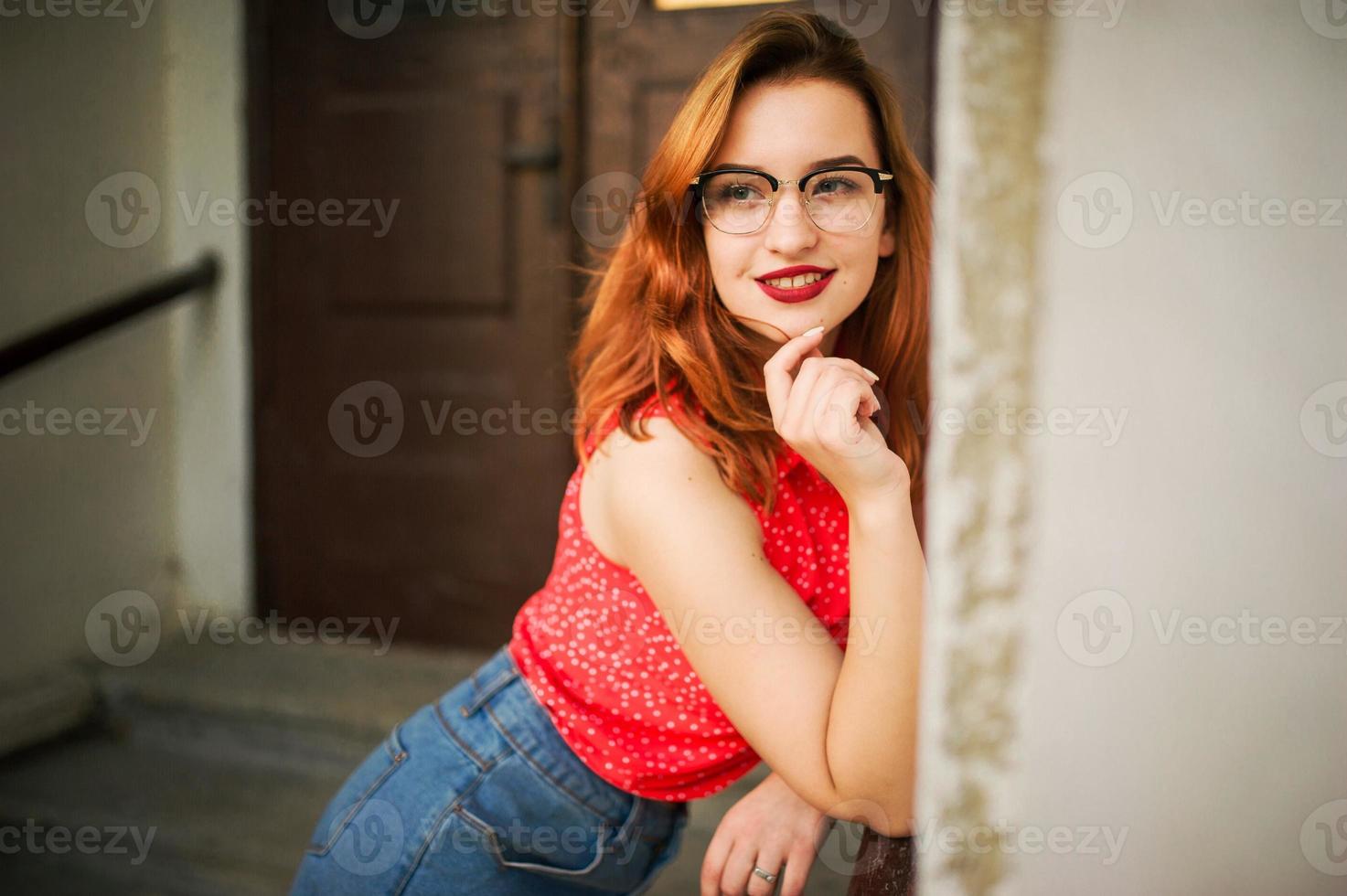 Attractive redhaired woman in eyeglasses, wear on red blouse and jeans skirt posing. photo